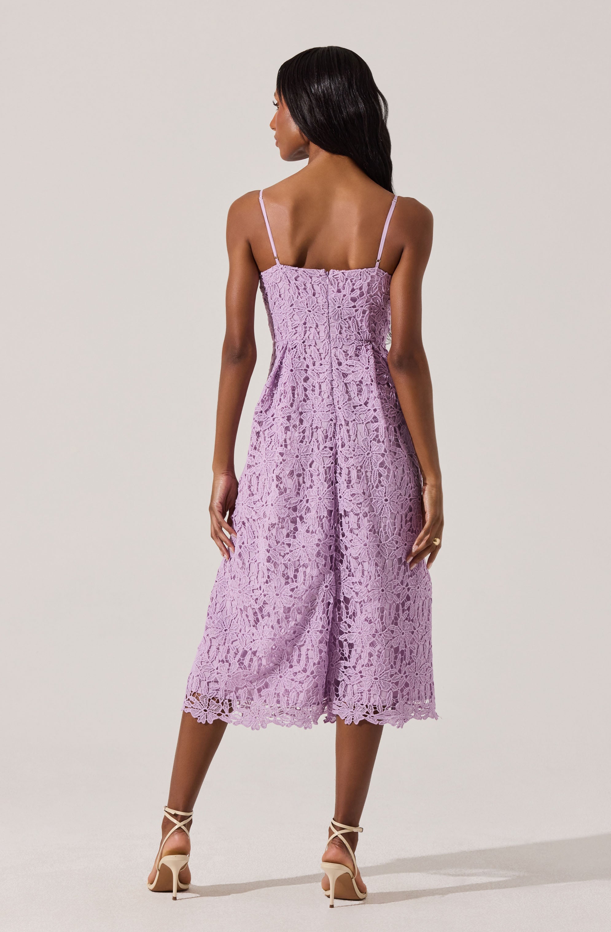 A woman is wearing a lavender-colored, sleeveless lace dress with thin spaghetti straps. The dress has a fitted bodice and a loose, midi-length skirt with a scalloped hem. She is also wearing strappy high-heeled sandals.