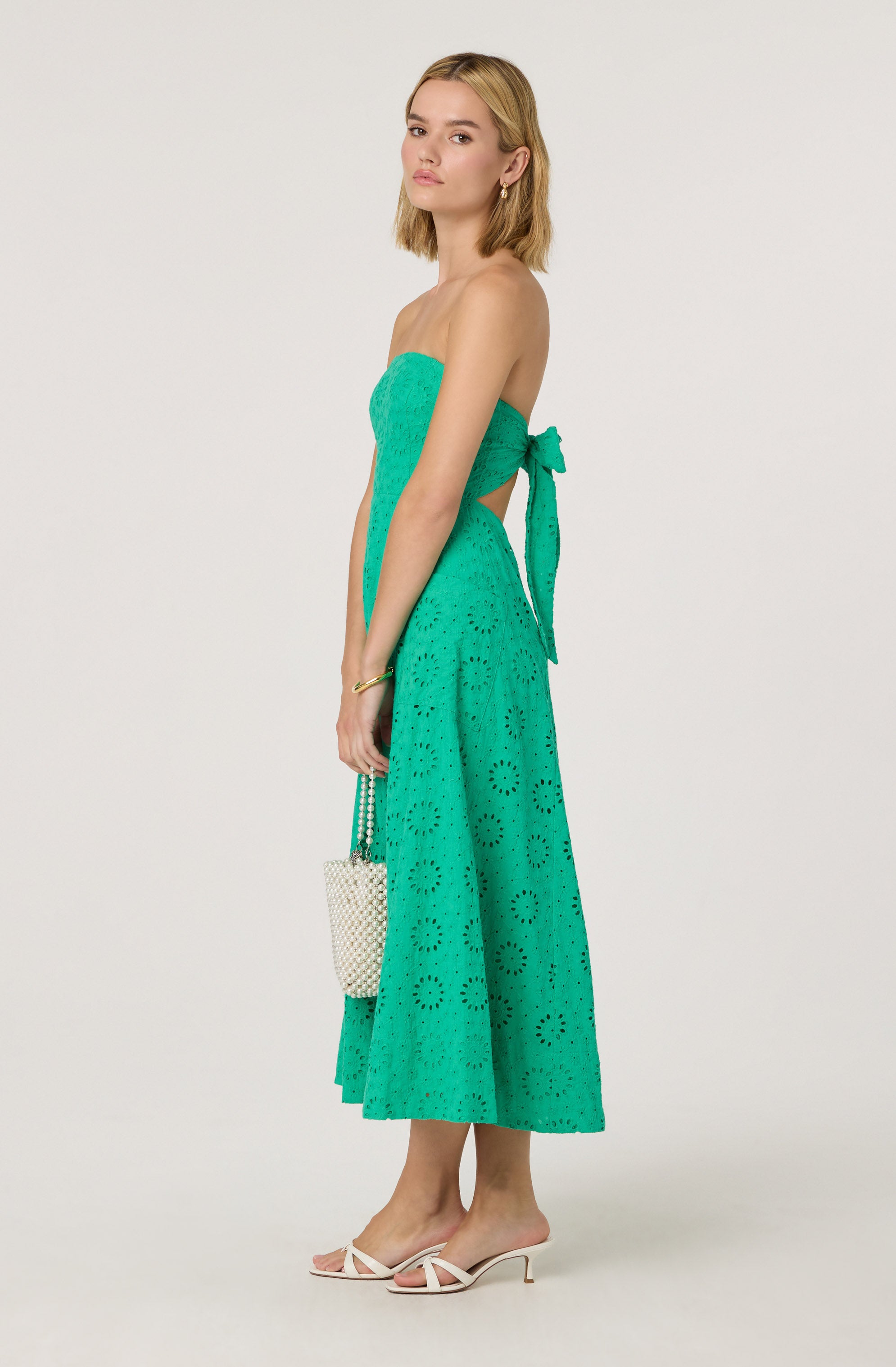 A woman is wearing a strapless, turquoise dress with eyelet embroidery. The dress has a fitted bodice and a high waist, with a matching sash tied in a bow on her left side. The skirt is mid-calf length, flowy, and features a circular eyelet pattern. She is also wearing white high-heeled sandals with thin straps.
