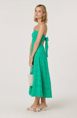A woman is wearing a strapless, turquoise dress with eyelet embroidery. The dress has a fitted bodice and a high waist, with a matching sash tied in a bow on her left side. The skirt is mid-calf length, flowy, and features a circular eyelet pattern. She is also wearing white high-heeled sandals with thin straps.