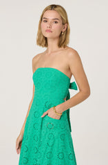 A young woman is wearing a strapless, bright turquoise dress with a subtle eyelet pattern throughout. The dress features a large pocket on the right side and a side bow detail at the waist. She accessorizes with gold hoop earrings and two gold bangles on her left wrist.