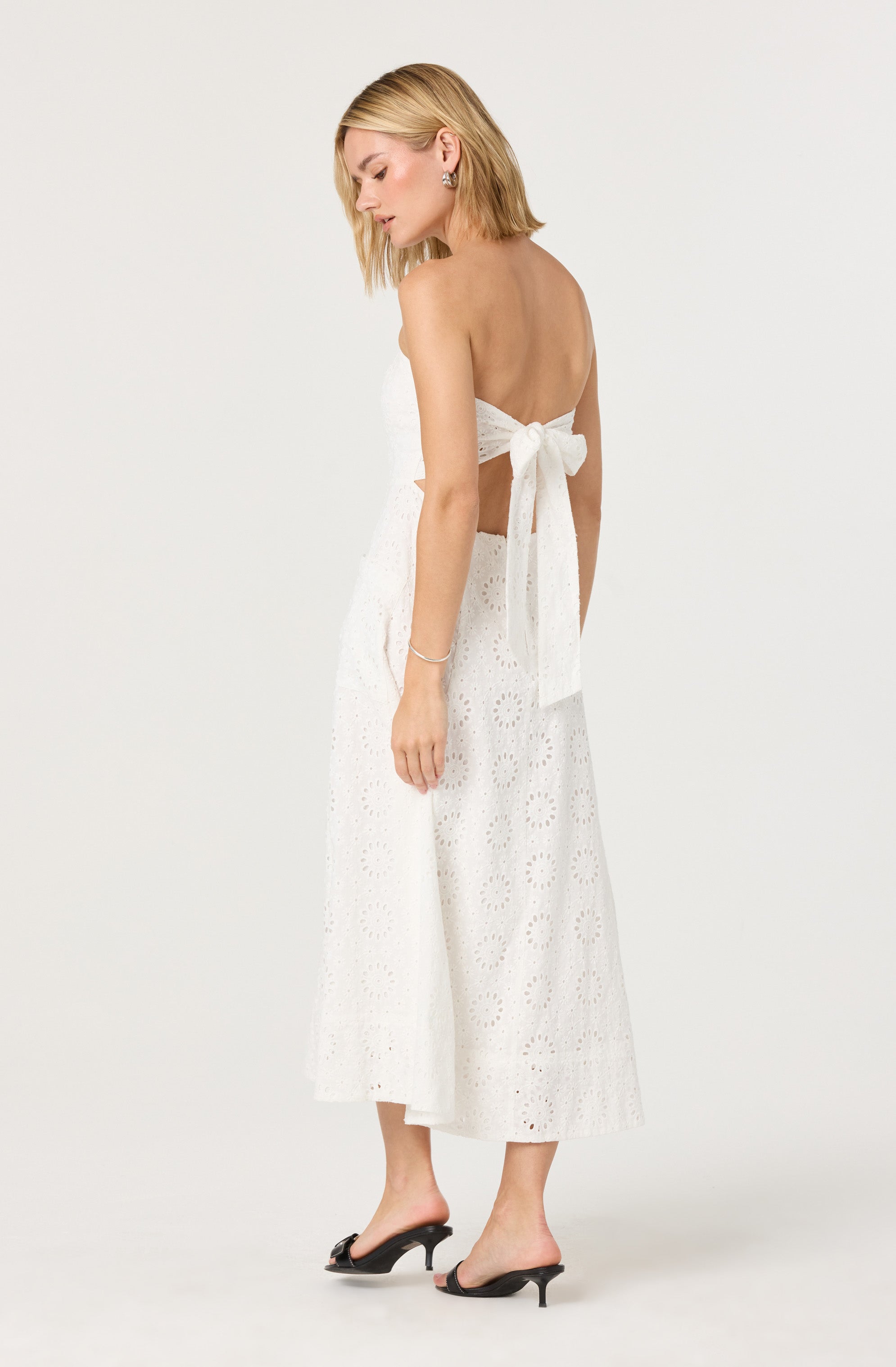 A woman is wearing a white, strapless midi dress with eyelet embroidery. The dress features a large bow tied at the back, and she is also wearing black open-toe high heels with a small heel.