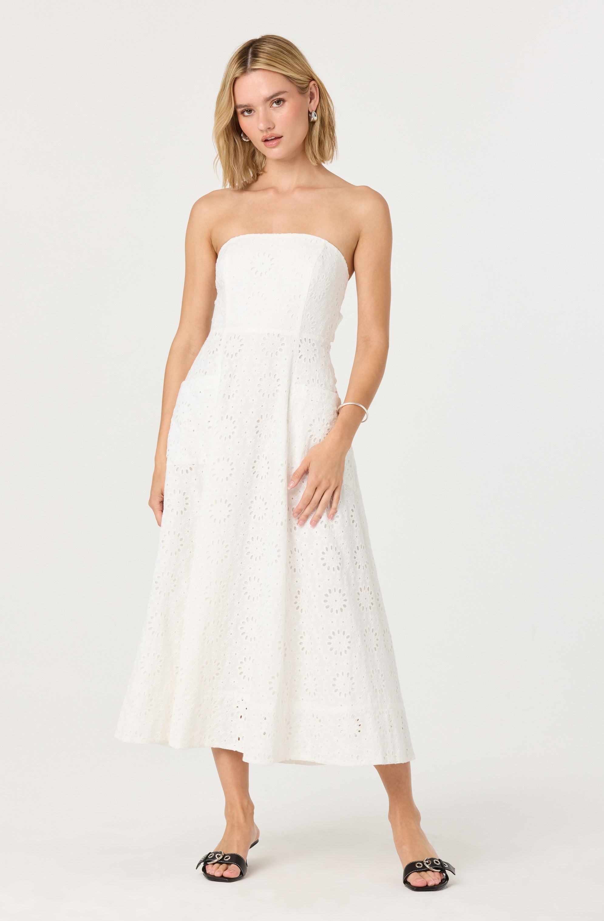 A woman is wearing a strapless, white, embroidered maxi dress with a fitted bodice and a flared skirt. The dress features intricate cut-out patterns throughout. She is also wearing open-toed black heels with decorative buckles, a bracelet, and earrings.