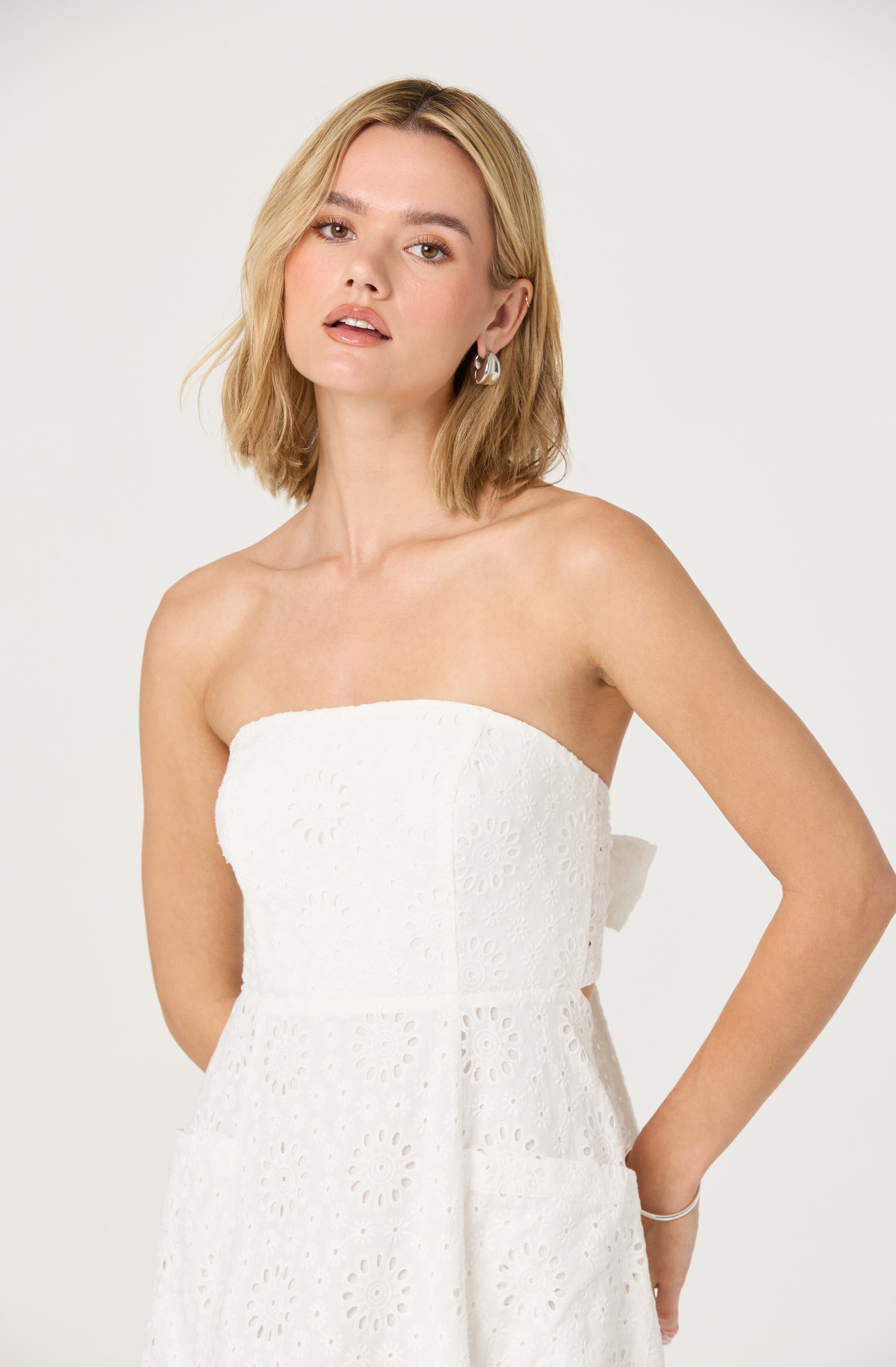 A woman is wearing a strapless white dress made of eyelet fabric, featuring circular cut-out patterns throughout. The dress has a fitted bodice and a slightly flared skirt.