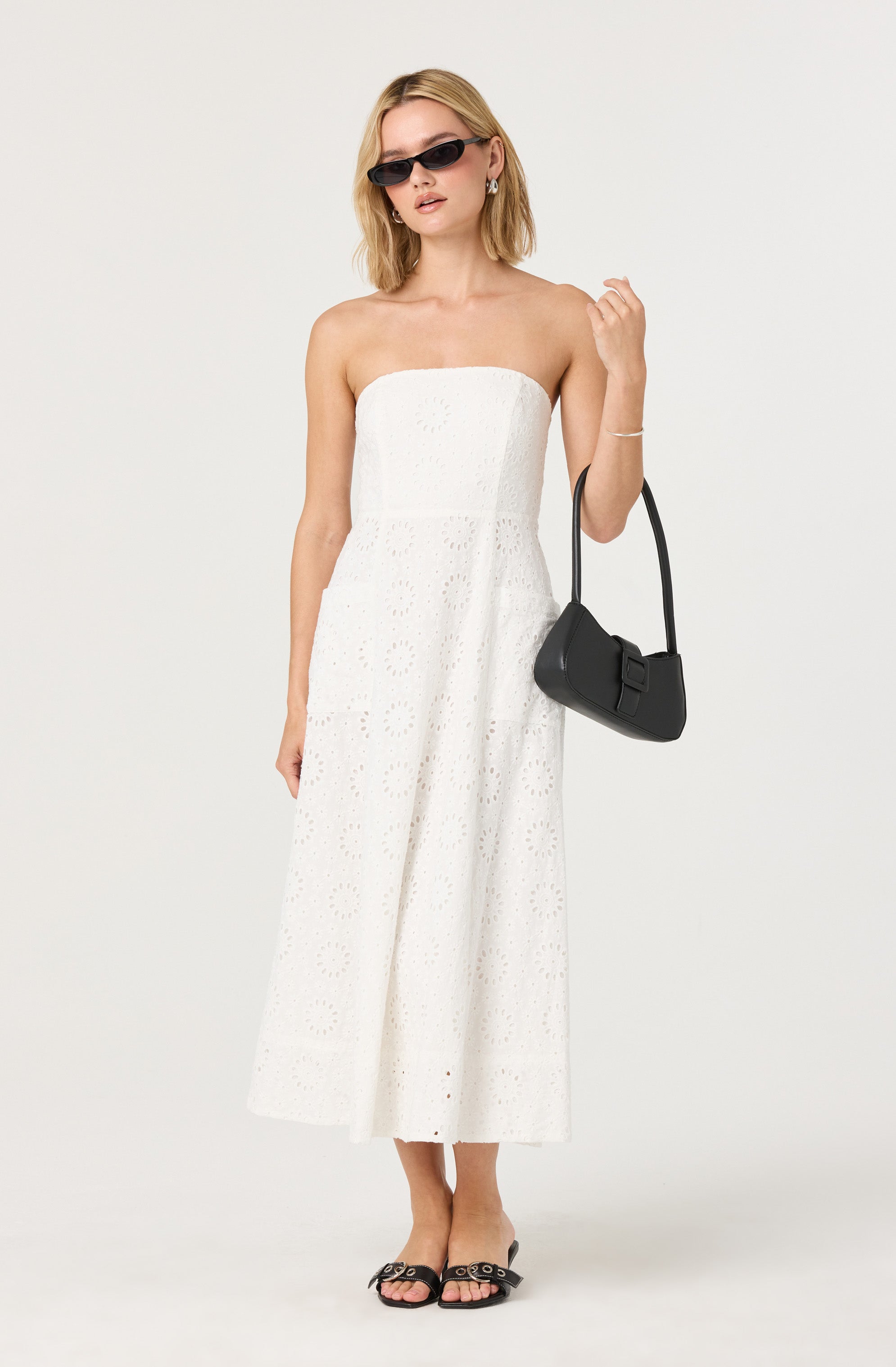A woman is wearing a strapless white dress with eyelet embroidery that features circular and floral cut-out patterns. She is accessorizing with black sunglasses, a small black shoulder bag with a bow detail, and black sandals with decorative metal rings. She has jewelry including earrings and a delicate bracelet.
