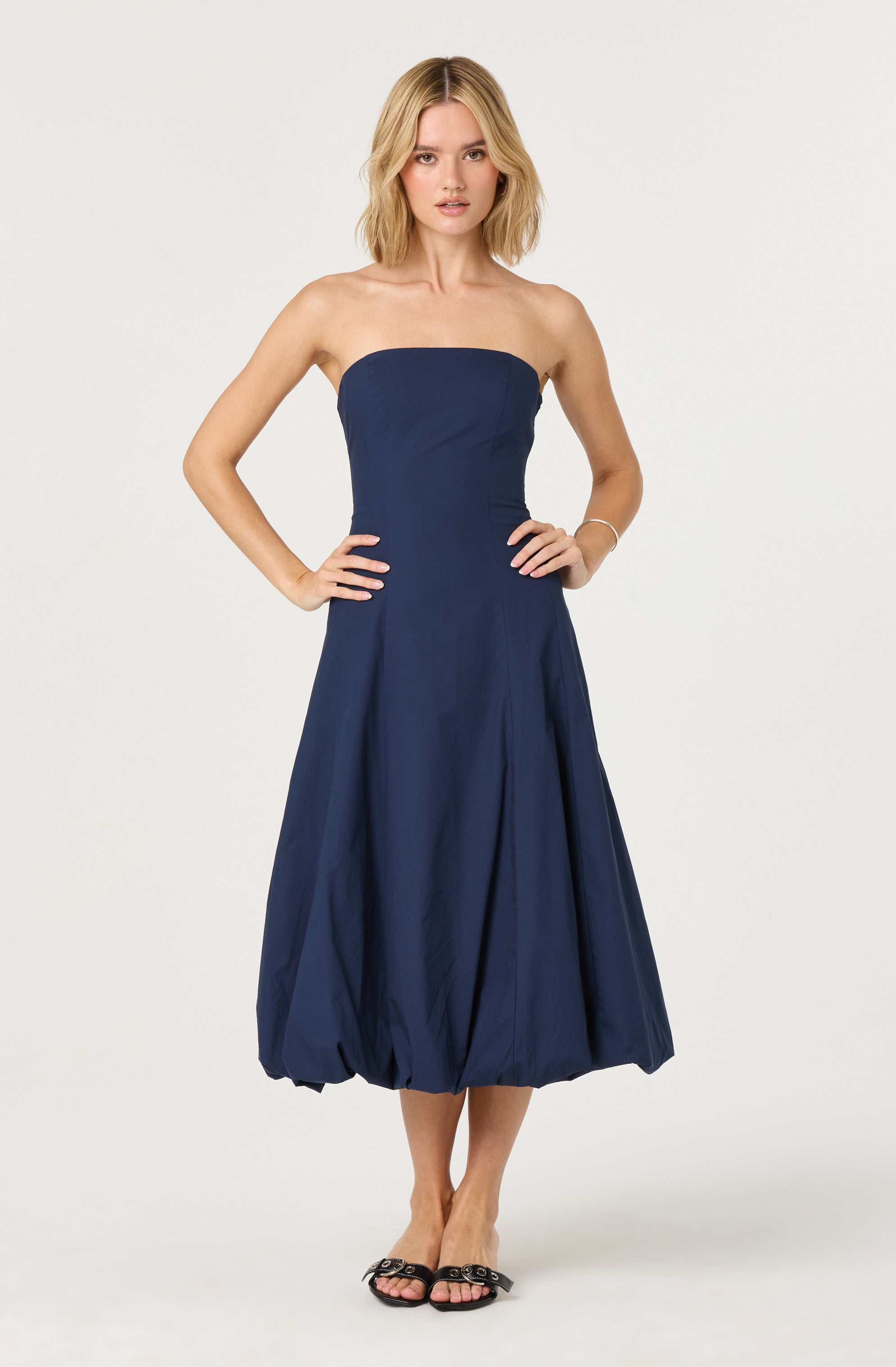A woman is wearing a strapless, navy blue dress with a fitted bodice and a voluminous, bubble-style skirt that falls below the knees. She is also wearing black open-toe sandals with decorative embellishments.