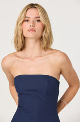 A woman is wearing a strapless navy blue dress that fits snugly around her torso. The dress has clean lines with visible seam details on the side. She accessorizes with a silver bracelet on her right wrist. Her blonde hair is styled in loose waves, and she has natural makeup.