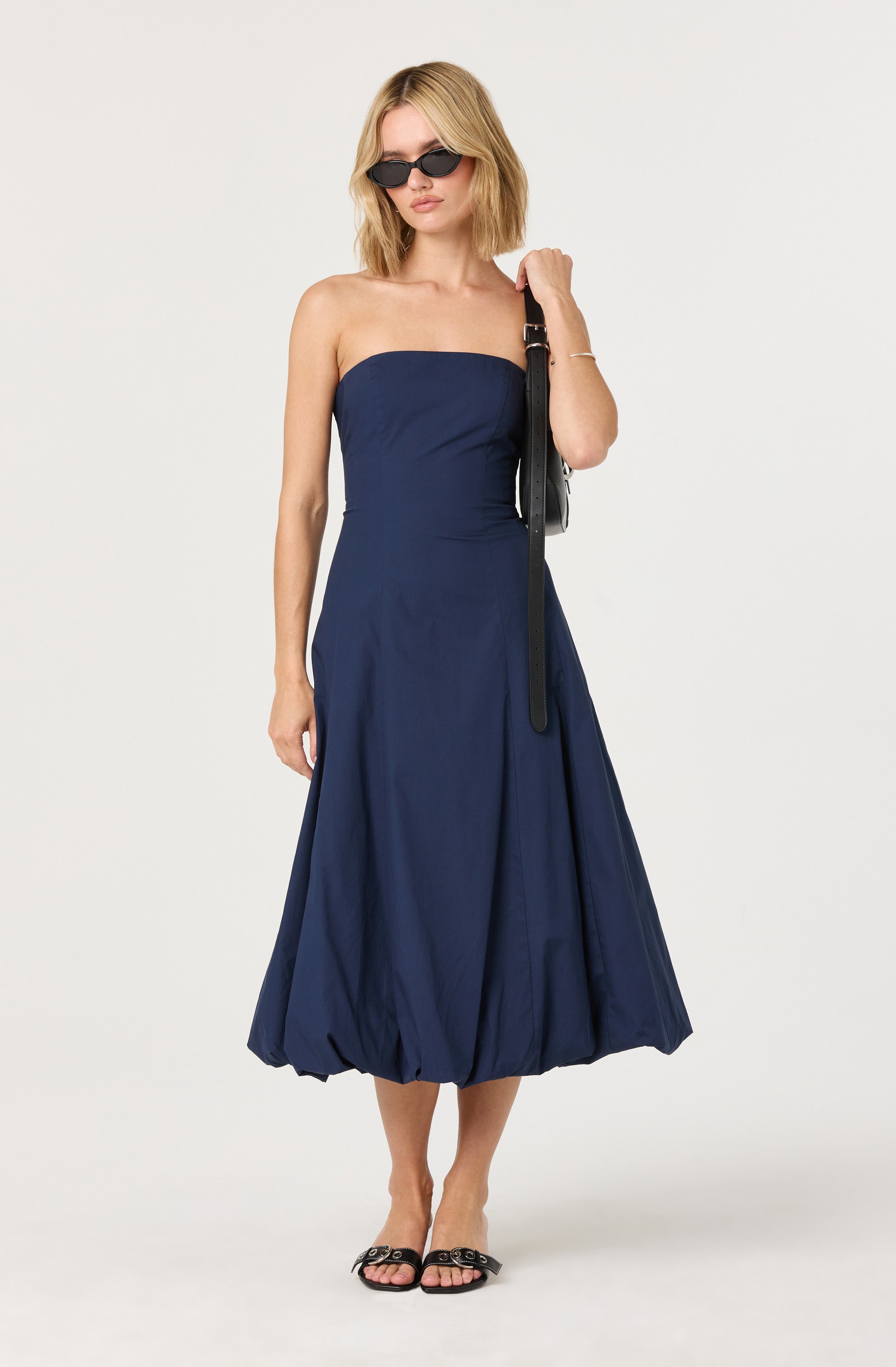A woman is wearing a strapless, dark blue, midi-length dress with a gathered, balloon-style hem. She accessorizes with black sunglasses, a silver bracelet, and black slide sandals with decorative embellishments on the straps. She is carrying a black shoulder bag with a long strap.