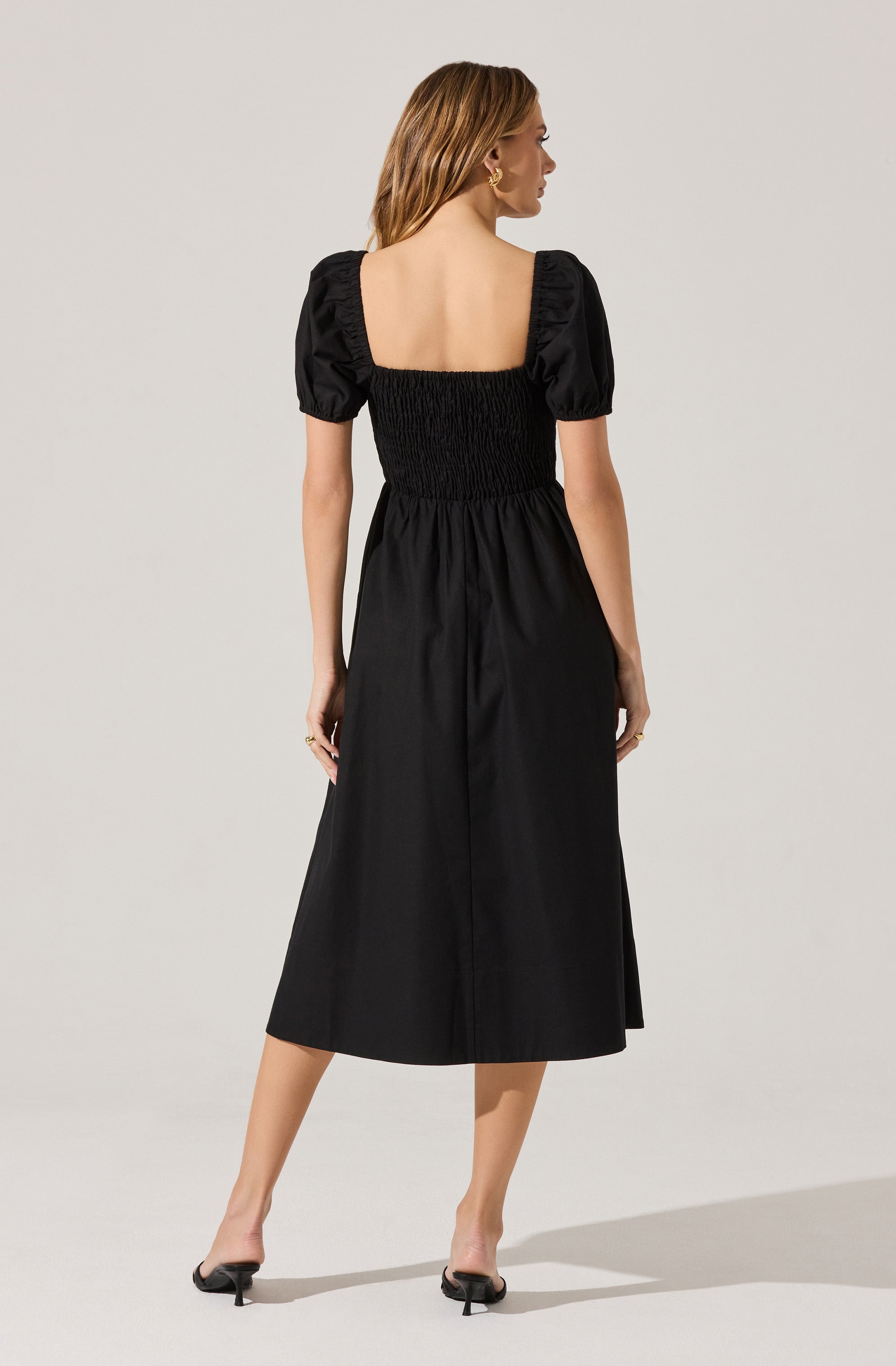 A woman is wearing a black dress with a fitted, elasticized waist and a square neckline. The dress features short, puffed sleeves with ruching detail. She is also wearing black open-toe heels.