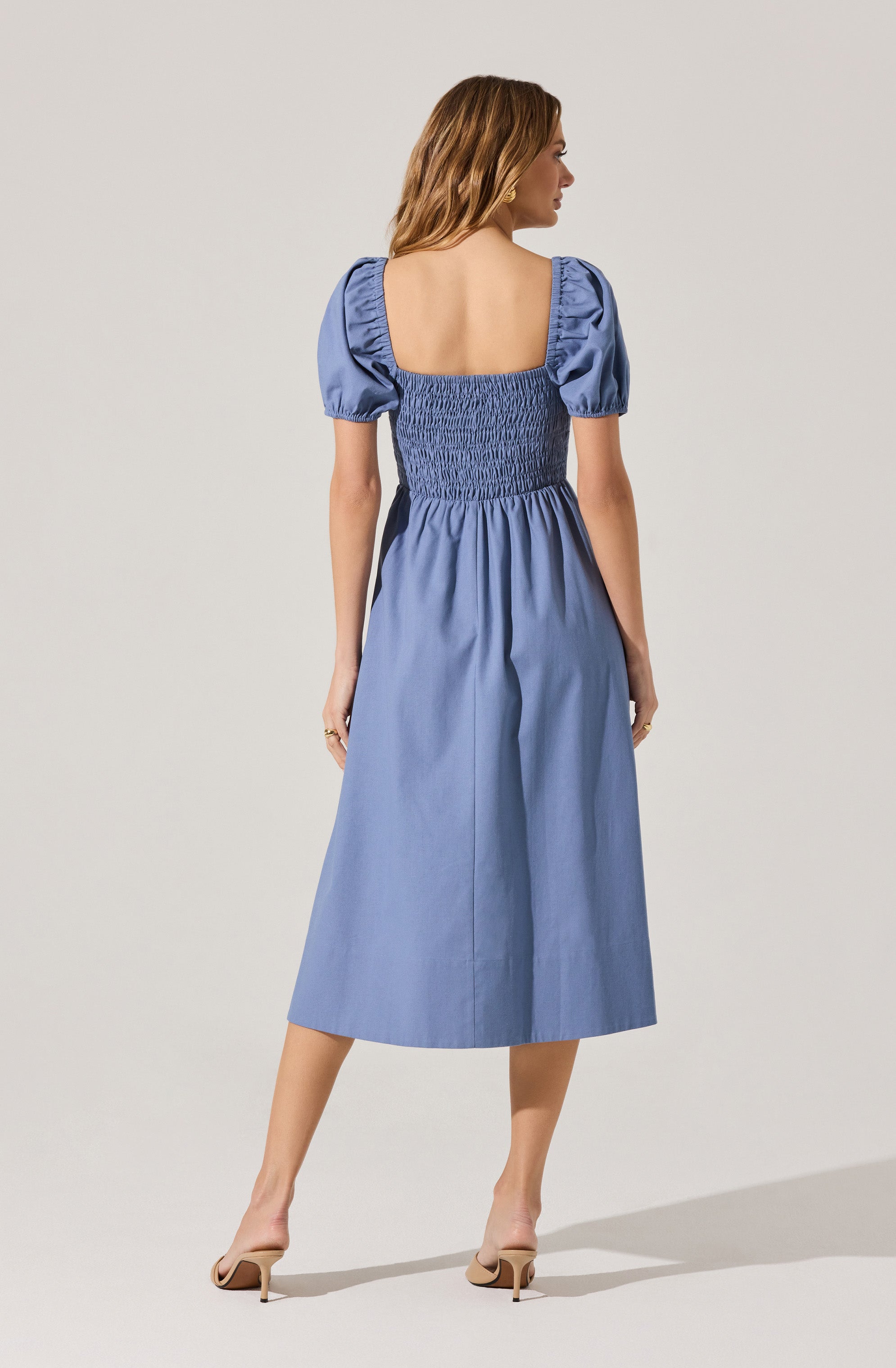 A woman is wearing a light blue midi dress with puffed short sleeves and a square neckline. The dress features a smocked bodice that extends to the waist, creating a fitted appearance, and the skirt is gathered, flowing loosely to mid-calf length. She is also wearing nude high-heeled shoes.