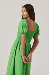 A woman is wearing a bright green dress with puffed short sleeves. The bodice of the dress features shirred or ruched detailing, creating a fitted waist, and the skirt is flowing and long. The dress has a wide, square neckline that exposes her shoulders and upper back.