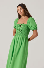 A woman is wearing a bright green dress with puffed short sleeves. The dress features a fitted, smocked bodice with a square neckline, decorated with a lace-up detail at the front. The skirt is long and flowing, made from a smooth fabric.
