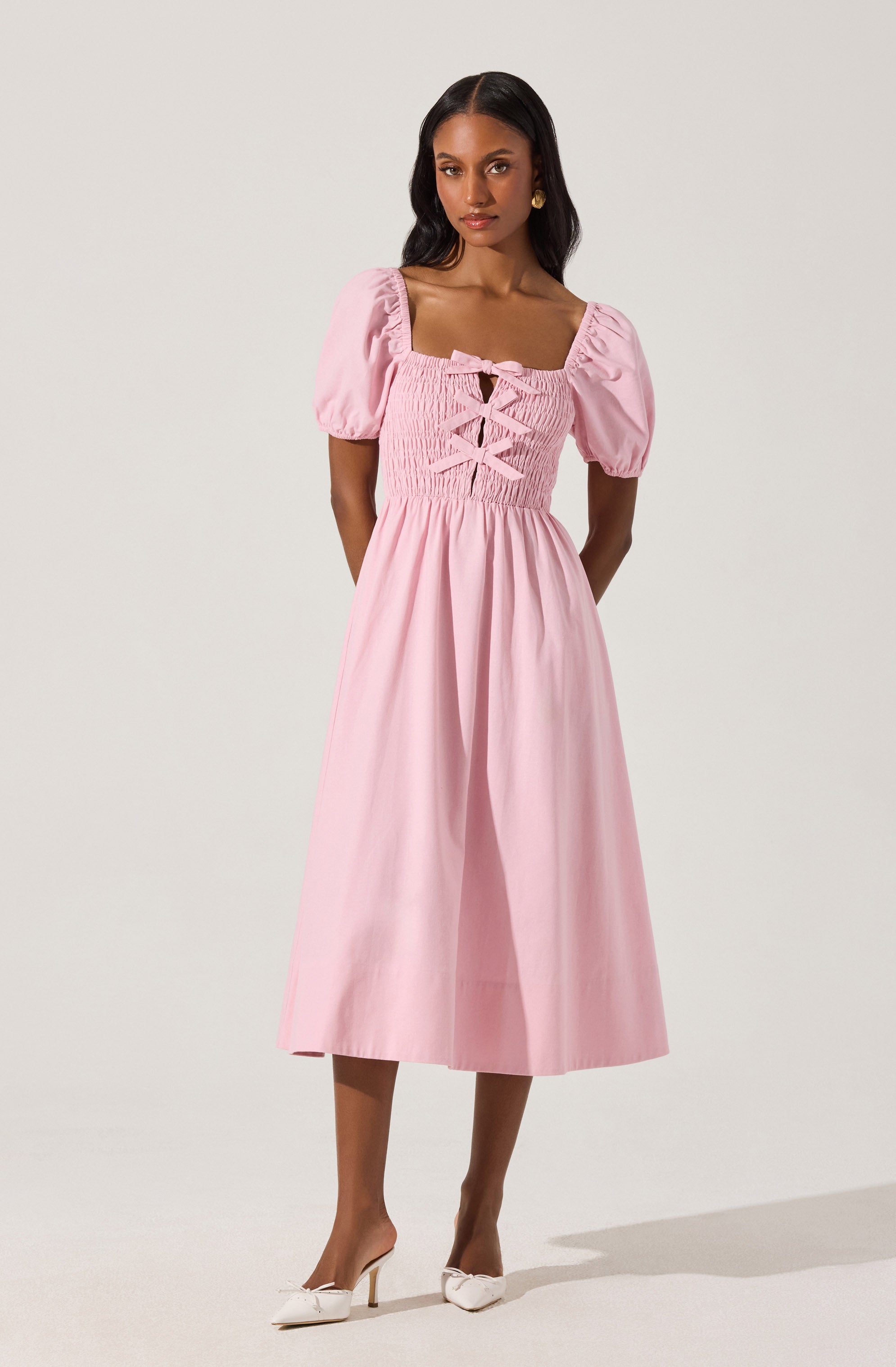 A woman is wearing a pink dress with puffed short sleeves, a square neckline, and a fitted bodice with a smocked design and lace-up detail at the front. The dress has a high waist and a full, midi-length skirt that falls below the knees. She is also wearing white pointed-toe heels.