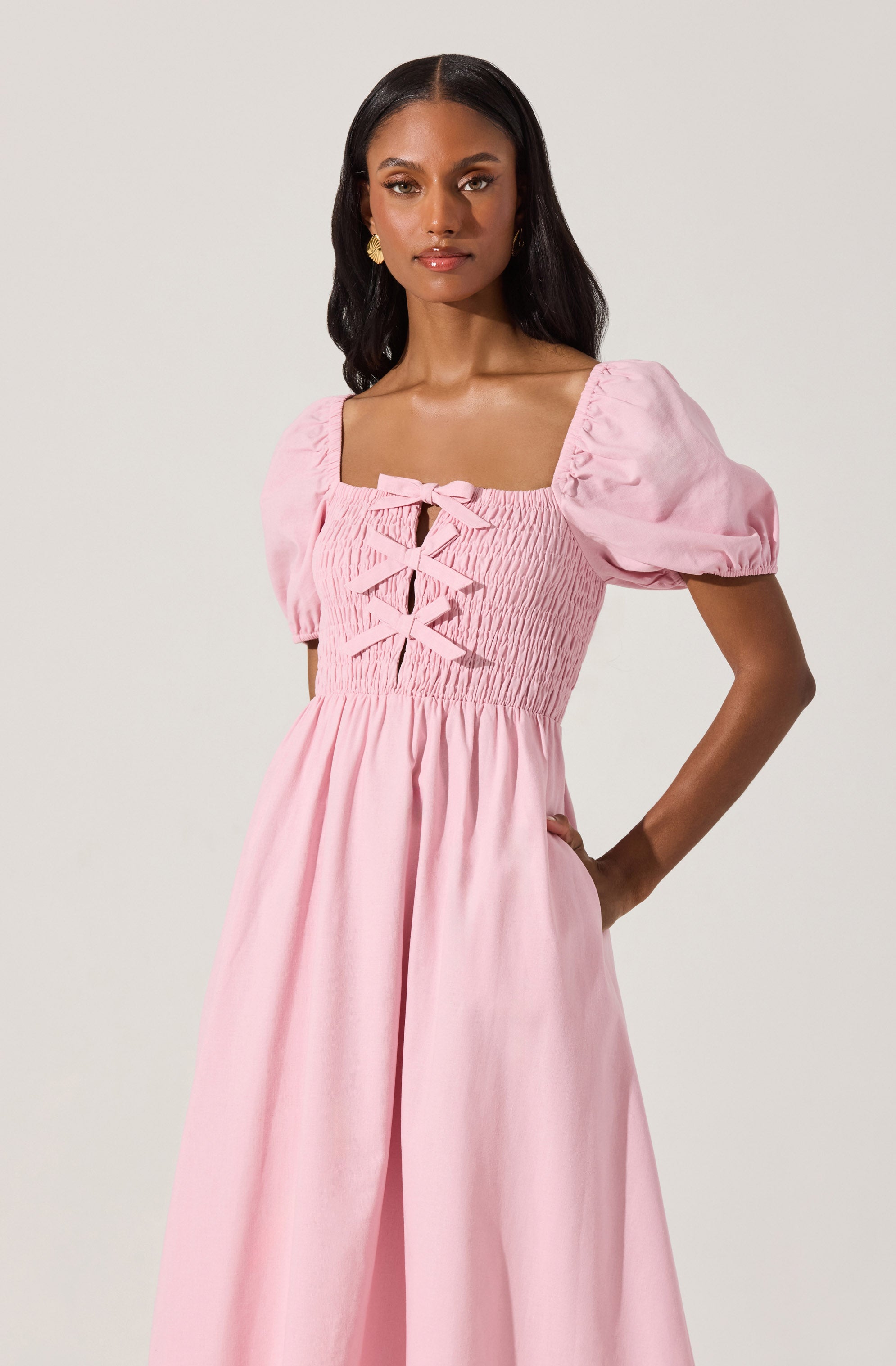 A woman is wearing a pink dress with puffed short sleeves, a square neckline, and a fitted bodice with smocking and decorative bows at the front. The skirt is long and flowing with a gathered waist.