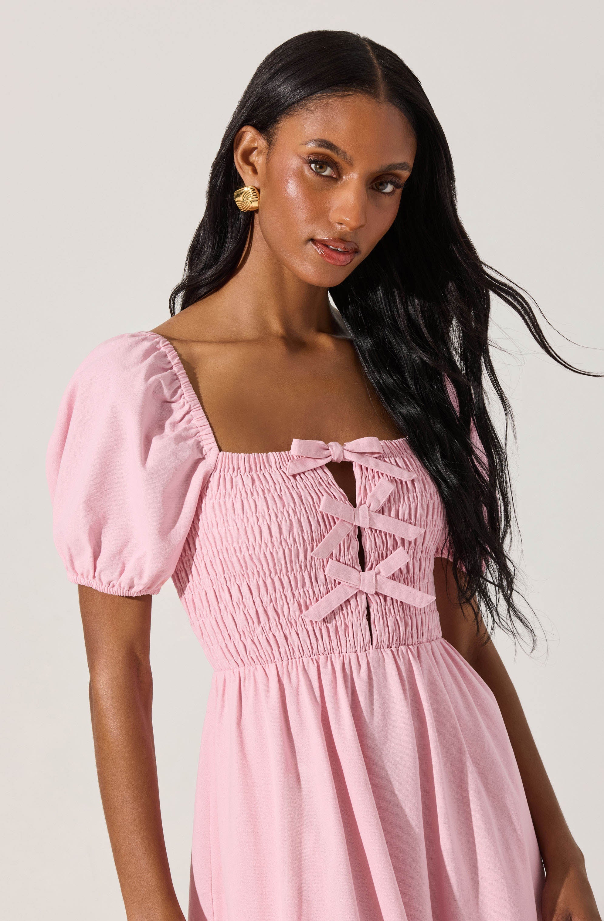A woman is wearing a pink dress with puffed short sleeves, a square neckline, and a smocked bodice. The bodice features decorative bows and a keyhole opening in the center.
