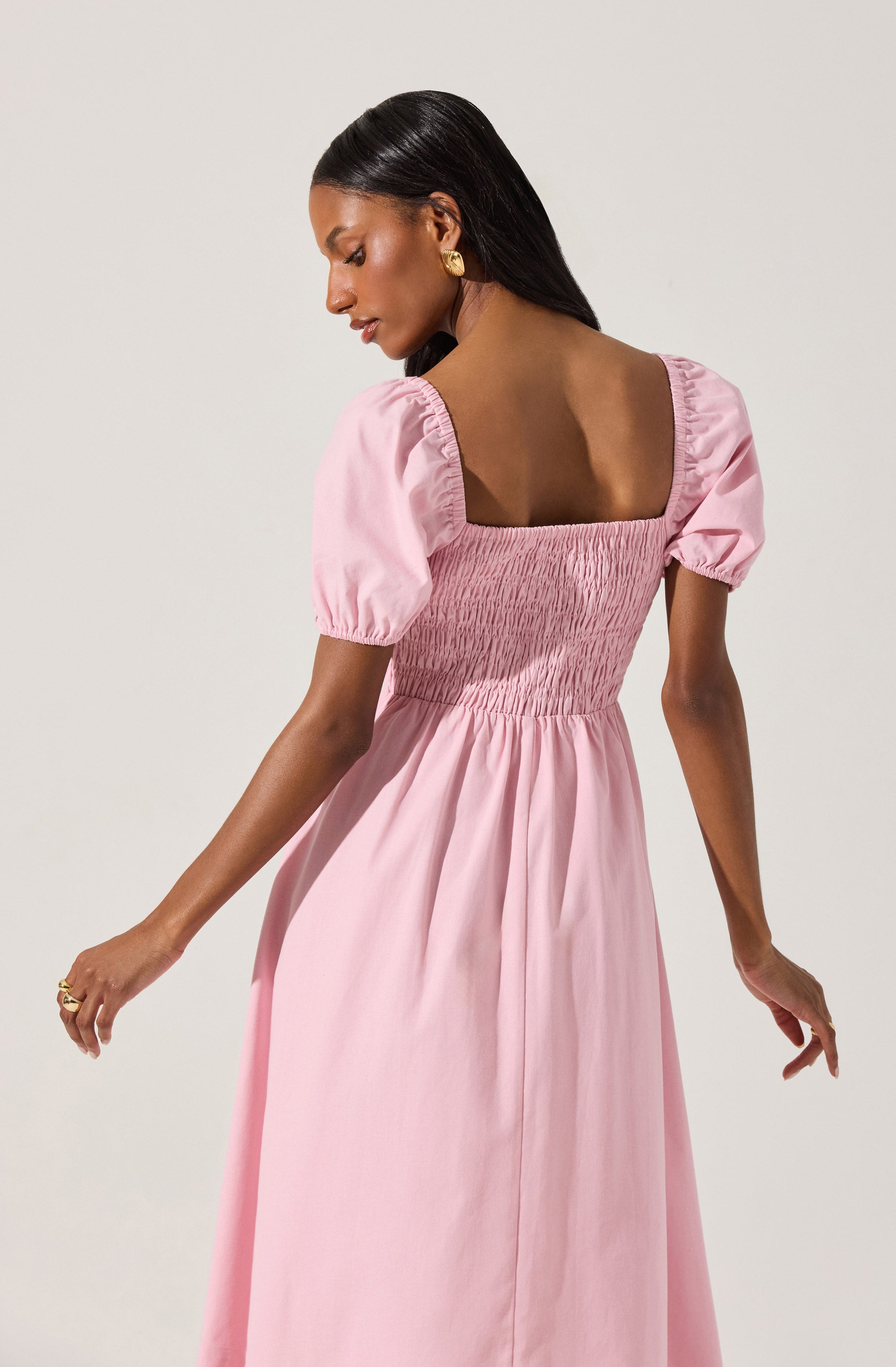 A woman is wearing a light pink, midi-length dress with puffed short sleeves. The bodice features shirred, gathered fabric that extends to the waist, creating a fitted look. The dress has a square neckline, and the skirt flows loosely from the waist down. The fabric appears lightweight and slightly shiny.
