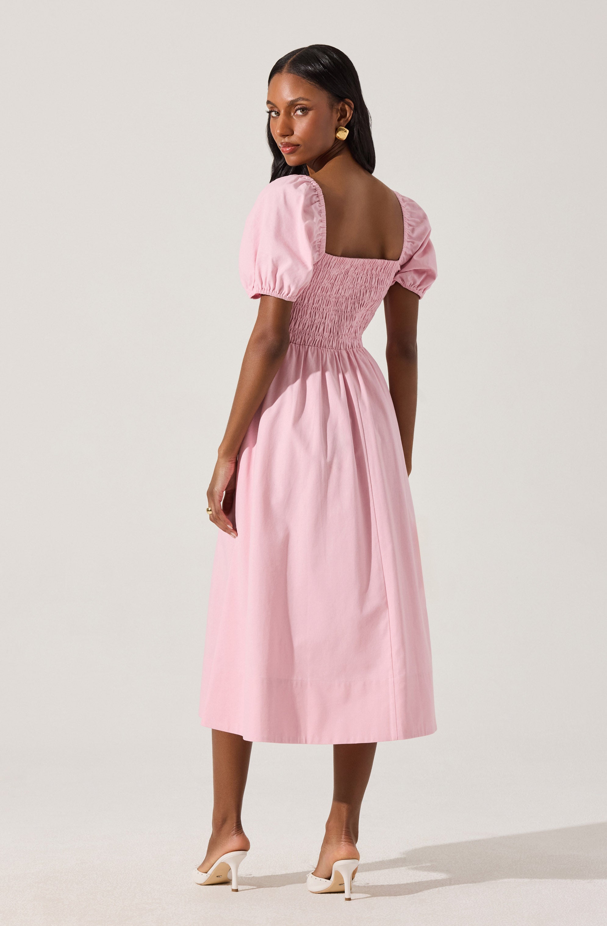 A woman is wearing a light pink dress with puffed short sleeves and a fitted, ruched bodice. The dress has a full, midi-length skirt that falls to her mid-calf. She is also wearing white high-heeled shoes with a pointed toe.