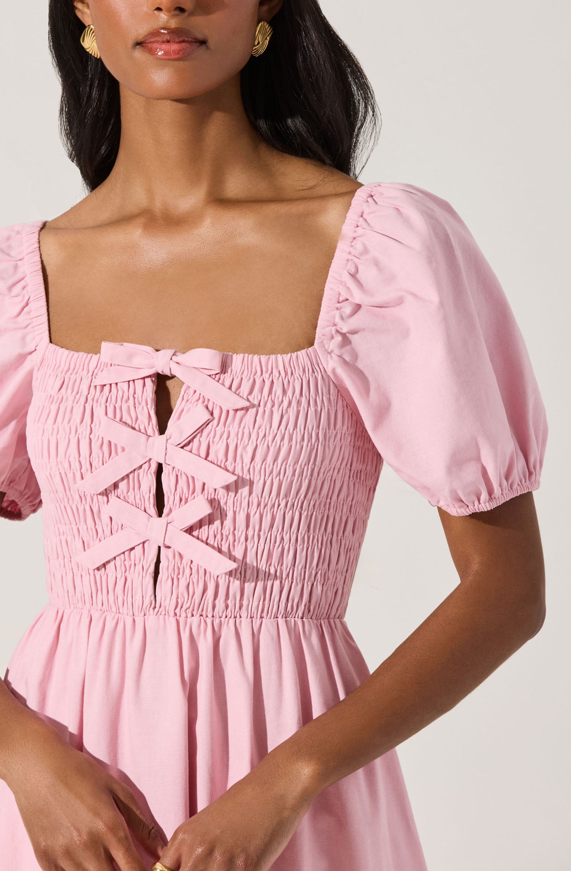 A woman is wearing a light pink dress with puffed short sleeves. The dress features a fitted bodice with shirred detailing and a decorative front with four horizontal bows laced through vertical slits. The skirt of the dress is gathered and flows loosely.