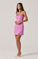 A woman is wearing a sleeveless, pink mini dress with spaghetti straps and small bow details at the top of the bust. The dress features a small slit on the side at the hem. She is also wearing beige pointed-toe high heels.