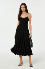 A woman is wearing a black, sleeveless midi dress with thin spaghetti straps and a fitted bodice. The dress has a flared skirt that falls below the knees. She is also holding a small black handbag with a gold accent, and wearing black open-toe high-heeled sandals.