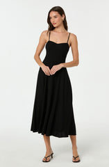 A woman is wearing a black sleeveless dress with thin spaghetti straps, a fitted bodice, and a flowing, ankle-length skirt. She is also wearing black high-heeled sandals.