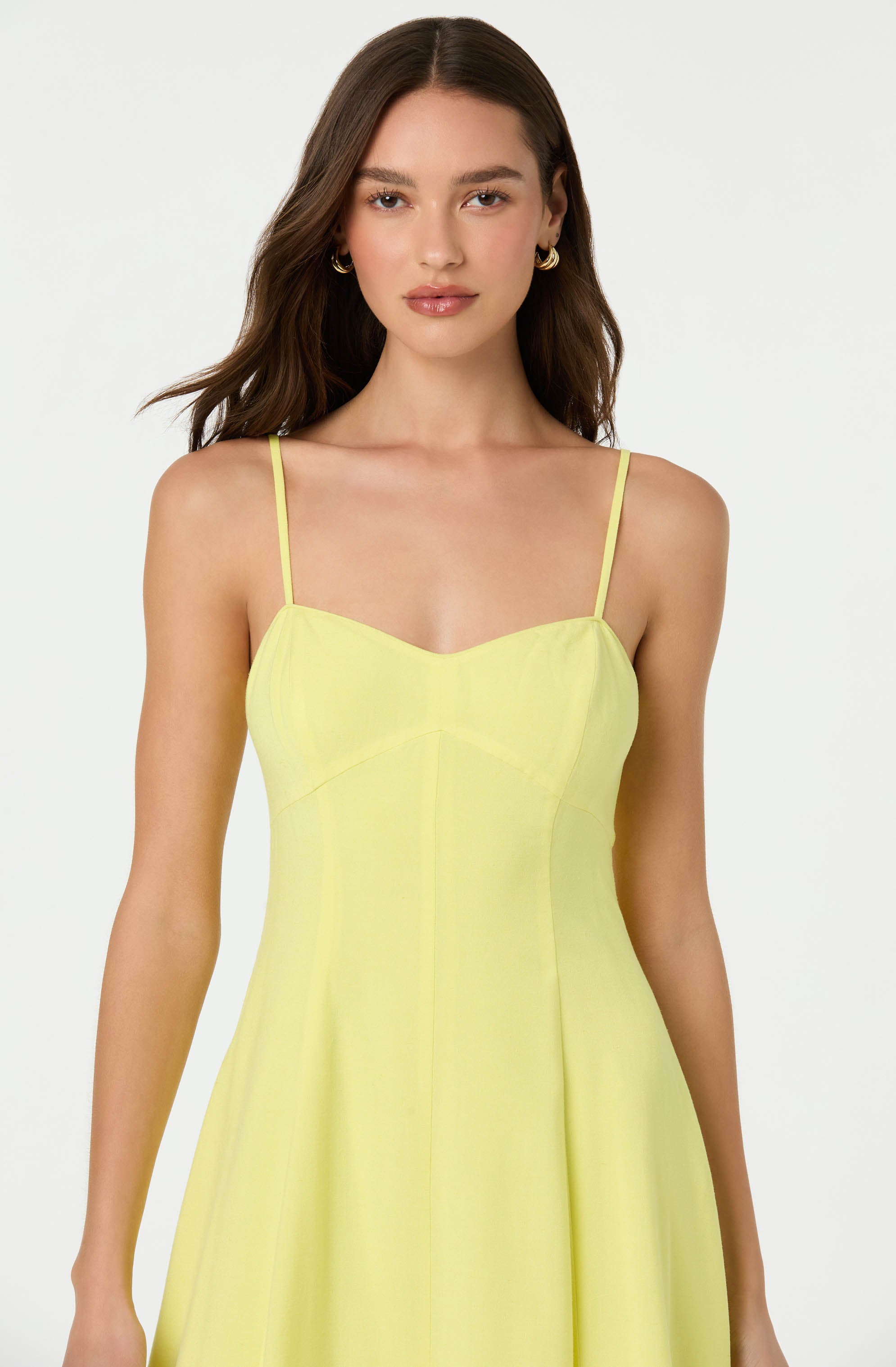 A woman is wearing a sleeveless, yellow dress with thin spaghetti straps. The dress features a fitted bodice with a sweetheart neckline and a slightly flared skirt.