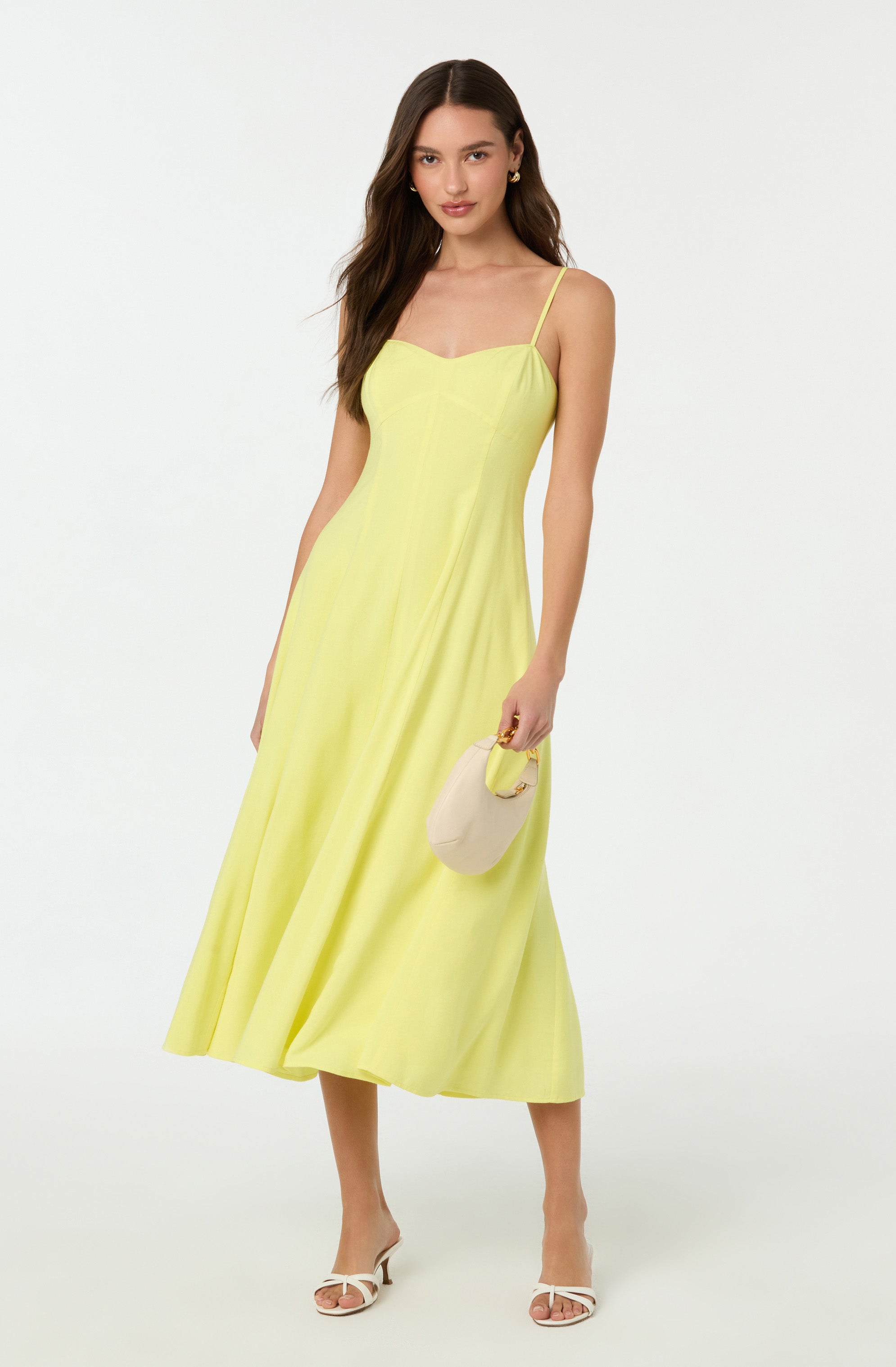 A woman is wearing a yellow sleeveless dress with thin spaghetti straps and a fitted bodice. The dress is long, flowing, and reaches down to her ankles. She is holding a light-colored, rounded handbag with a yellow handle in her right hand. She is also wearing white open-toe high heels with a slim strap across the toes.