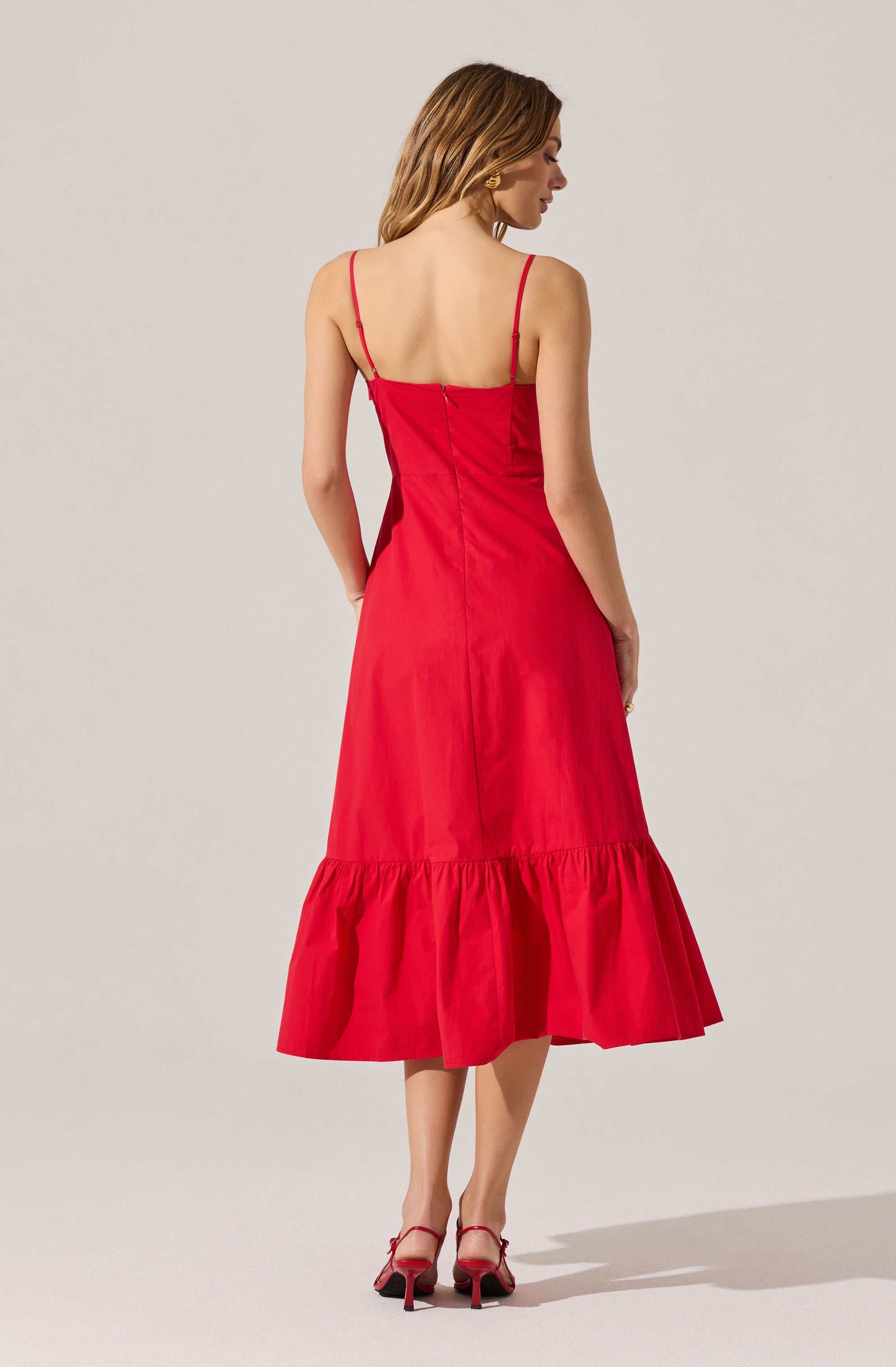 A woman is wearing a sleeveless, red midi dress with thin spaghetti straps and a fitted bodice. The dress features a gathered ruffle hem that falls below the knees. She pairs the outfit with red high-heeled sandals that have ankle straps.
