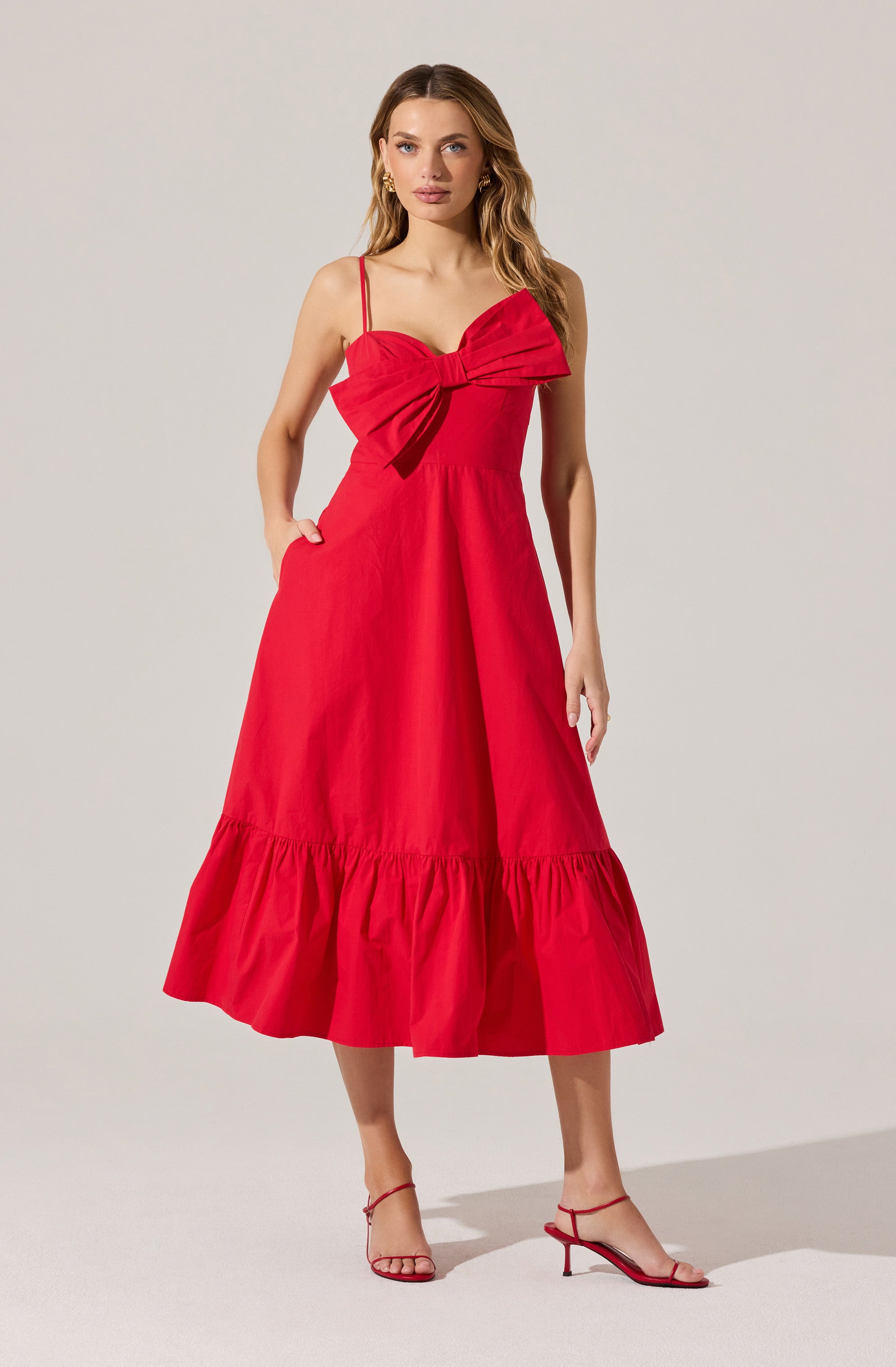 A woman is wearing a sleeveless red dress with thin spaghetti straps. The dress features a large bow detail at the bust area, and has a fitted waist. The skirt is midi length with a tiered ruffle at the hem. She is also wearing red high-heeled sandals with thin straps.