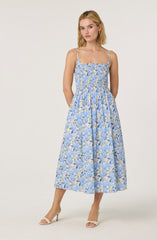 A woman is wearing a sleeveless, ankle-length dress with thin spaghetti straps. The dress has a fitted, smocked bodice and a full, flowing skirt. The fabric features a light blue and beige floral pattern. She is also wearing beige open-toe heels.