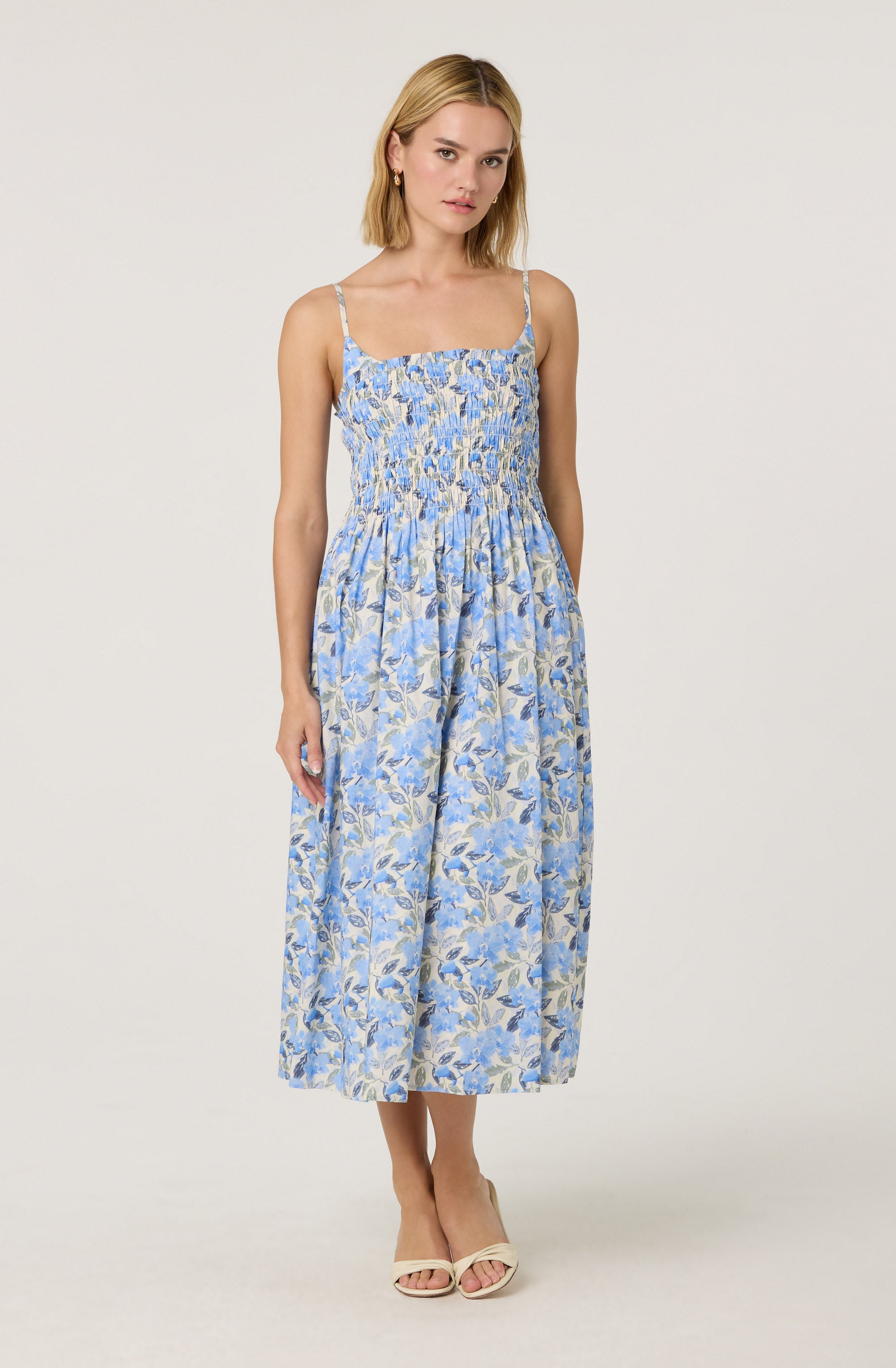 A woman is wearing a sleeveless, ankle-length dress with thin spaghetti straps. The dress features a fitted, smocked bodice and a flowing skirt with a floral pattern in shades of blue, beige, and light lavender. She is also wearing open-toed, light-colored high-heeled sandals.