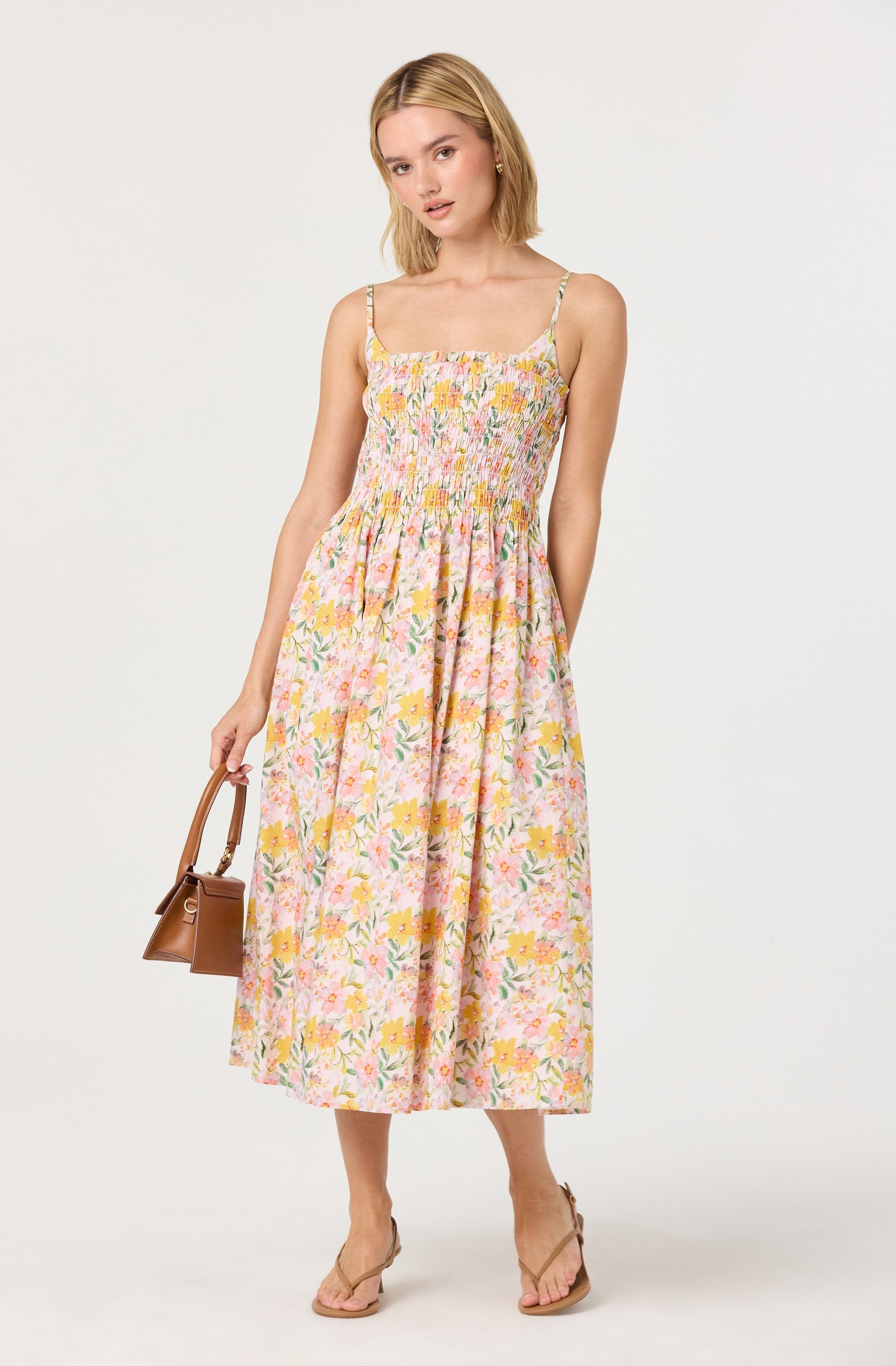 A woman is wearing a sleeveless, ankle-length summer dress with a floral pattern featuring pink, yellow, and green colors. The dress has thin spaghetti straps and a smocked bodice. She is holding a small brown handbag in her right hand and is wearing beige sandals.