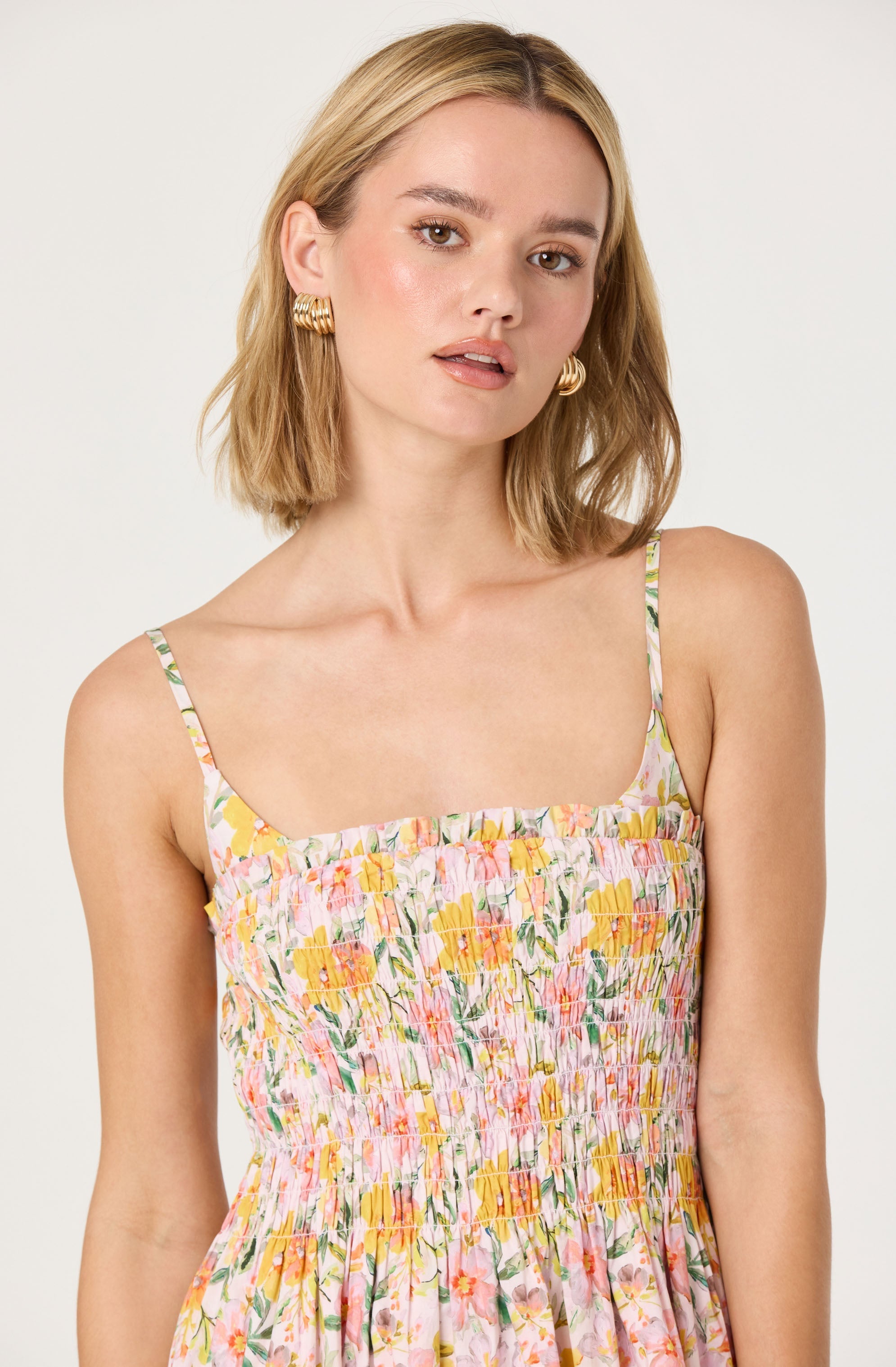 A woman is wearing a sleeveless dress with thin spaghetti straps. The dress features a floral print with colors including yellow, pink, green, and white. The fabric has a gathered, smocked texture around the bodice, creating a fitted look, and the skirt portion is loose and flowing.