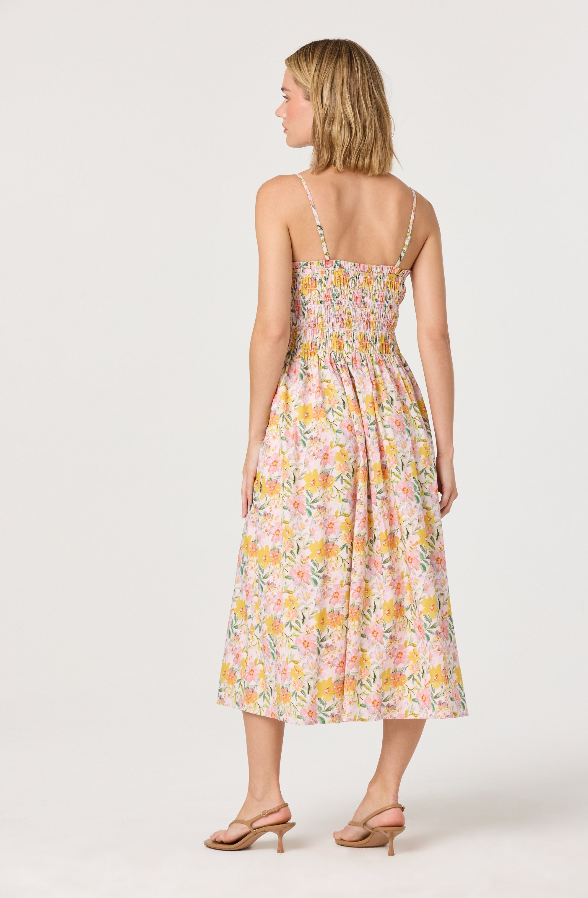 A woman is wearing a sleeveless sundress with thin, spaghetti straps. The dress features a smocked bodice and a flowy, midi-length skirt with a colorful floral pattern in shades of pink, yellow, green, and white. She is also wearing nude high-heeled sandals.