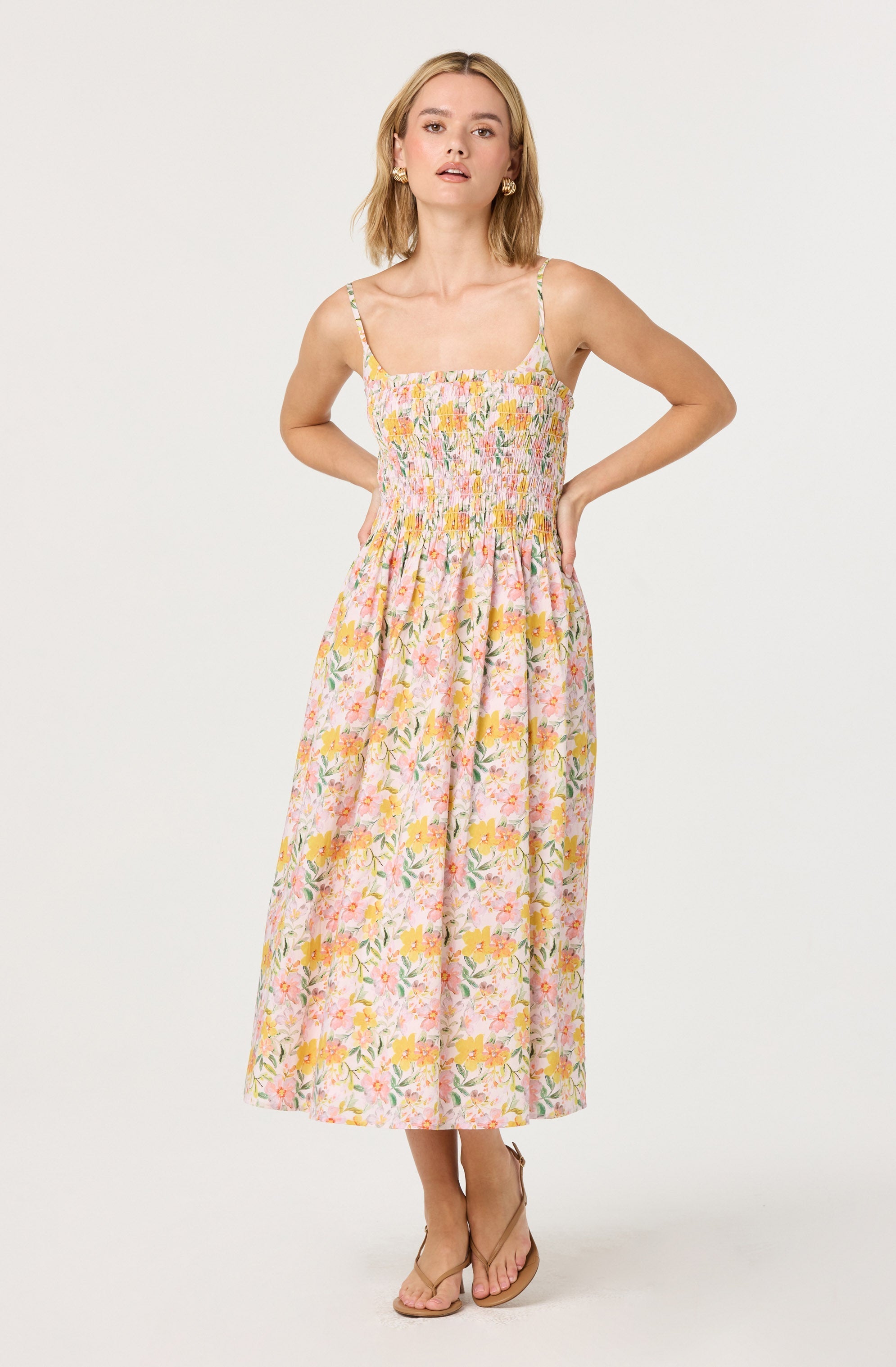 A woman is wearing a sleeveless, spaghetti strap, ankle-length dress with a floral pattern in shades of pink, yellow, green, and white. The dress has a smocked bodice and a gathered skirt, creating a casual, summery look. She is wearing nude-colored sandals.