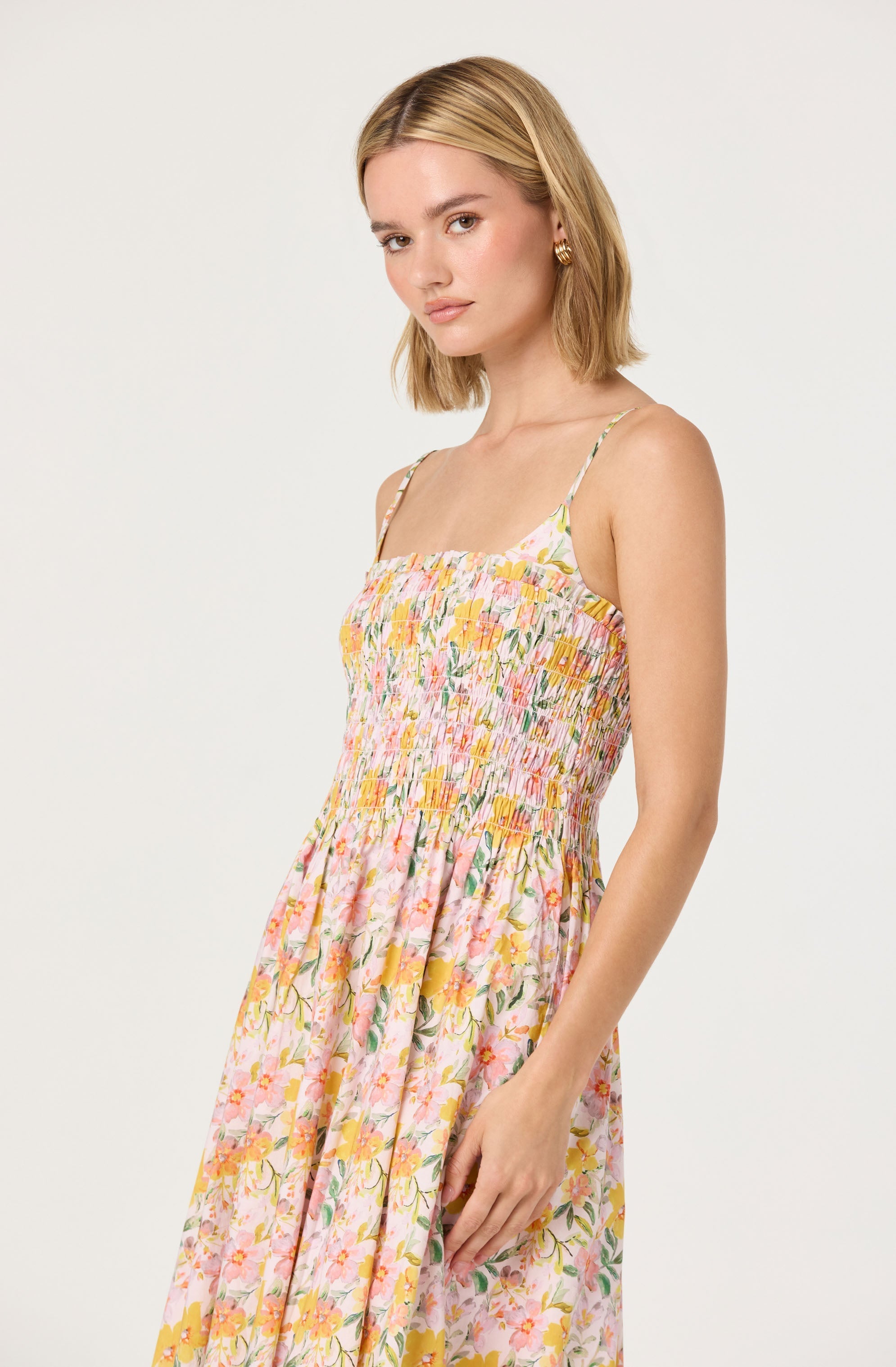 A young woman is wearing a sleeveless, floral summer dress with spaghetti straps. The dress features a fitted, smocked bodice with multicolored floral patterns, and a flowing skirt that extends below the waist. The floral print is predominantly in shades of pink, yellow, and green on a light-colored background.