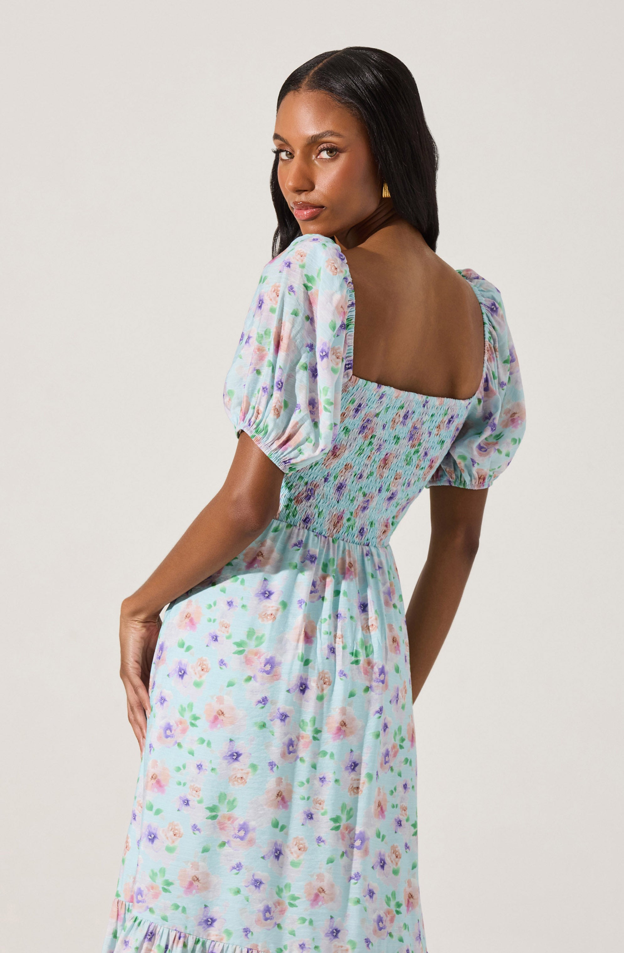 A woman is wearing a light-colored dress with a floral pattern featuring pastel pink, purple, and green flowers. The dress has puffed short sleeves and a square neckline. The bodice is smocked with a multicolored pattern, and the skirt flows down with a loose fit.