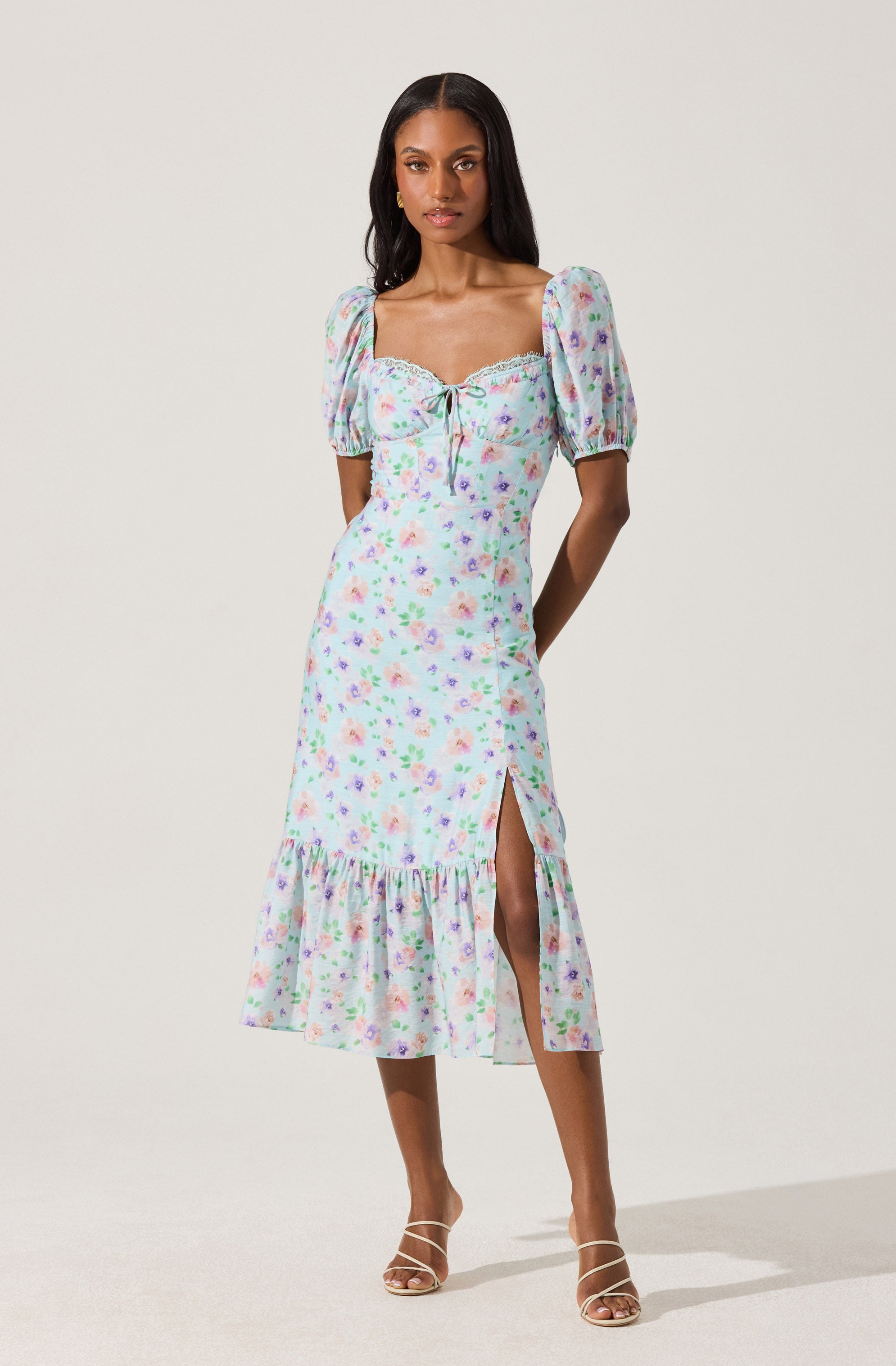 A woman is wearing a midi dress with a pastel floral print. The dress has puffed short sleeves, a square neckline with a lace trim, and a front tie detail at the bust. It features a high slit on one side and a ruffled hem at the bottom, adding a feminine touch. The fabric appears lightweight and flowy. She is paired with strappy high-heeled sandals.