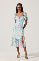 A woman is wearing a midi dress with a pastel floral print. The dress has puffed short sleeves, a square neckline with a lace trim, and a front tie detail at the bust. It features a high slit on one side and a ruffled hem at the bottom, adding a feminine touch. The fabric appears lightweight and flowy. She is paired with strappy high-heeled sandals.