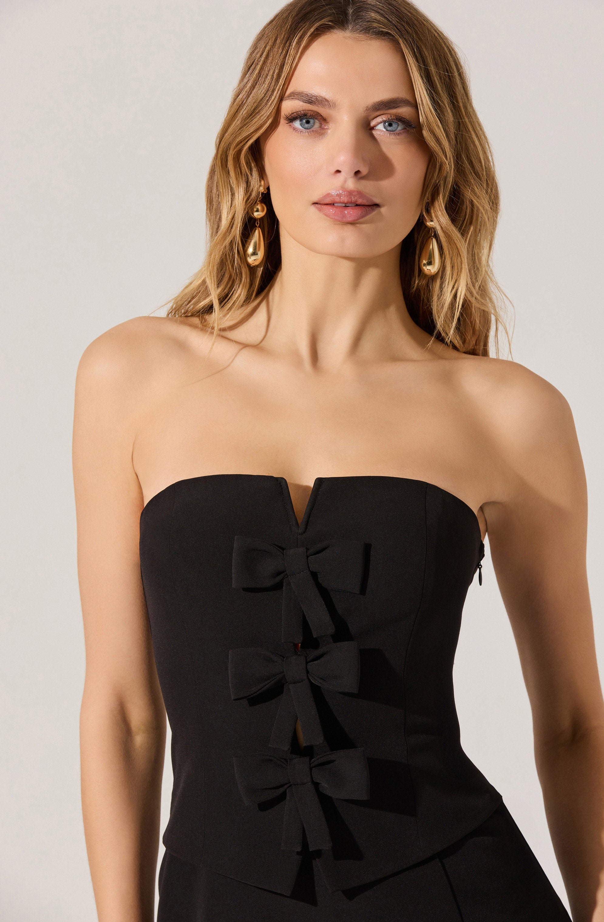 A woman is wearing a strapless black top with a sweetheart neckline. The top features three decorative bows running vertically down the center. She is accessorized with large, gold, teardrop-shaped earrings.