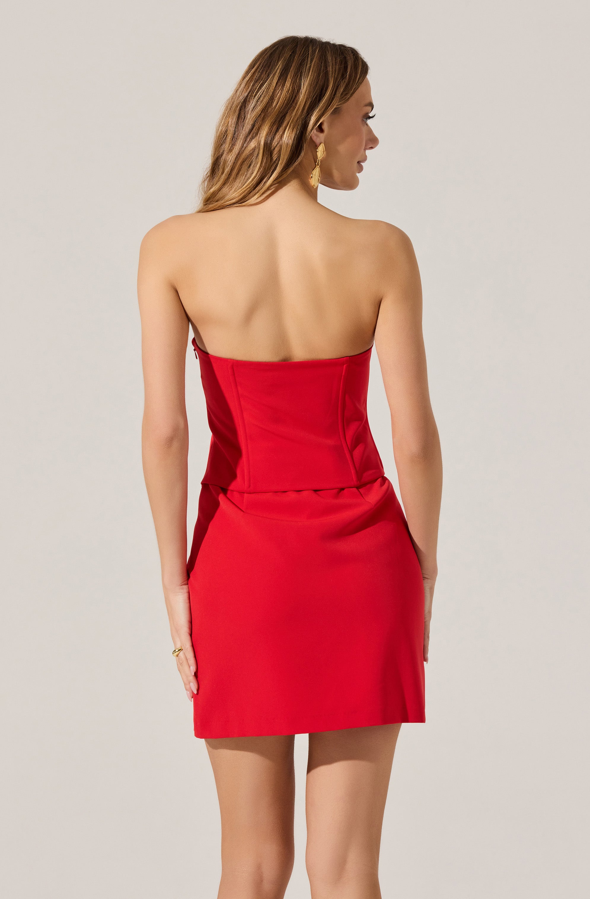 A woman is wearing a strapless, fitted red dress that is knee-length. The dress has a smooth, slightly shiny fabric and features seam details that run down the sides. The back of the dress is plain, with no visible embellishments or patterns.