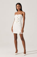 A woman is wearing a strapless, white mini dress with a fitted bodice adorned with three decorative bows down the center. The dress has a small slit on the left side near the hem. She is accessorizing with large, round earrings and white shoes with a bow detail.