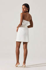 A woman is wearing a strapless, fitted white dress that reaches above the knees. She has gold earrings and is wearing light-colored high-heeled shoes. The dress features a straight neckline and a smooth, sleek fabric.