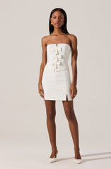 A woman is wearing a strapless white dress with a fitted bodice featuring three decorative bows down the front. The dress is knee-length with a small slit on the front left side. She is also wearing white pointed-toe high heels with a bow detail.