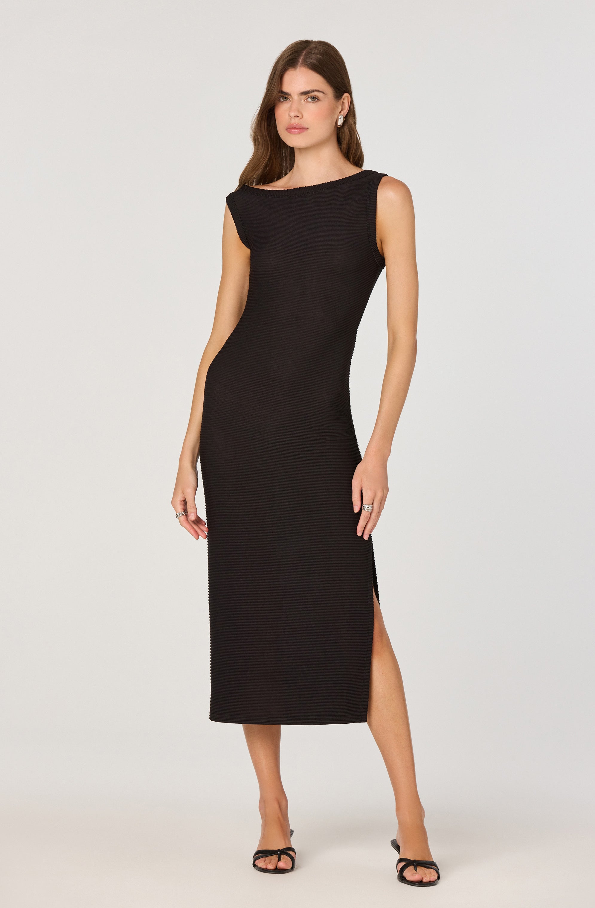 A woman is wearing a sleeveless, black, full-length dress with a high slit on the side. The dress has a boat neckline and a fitted silhouette. She is also wearing black open-toe high-heeled sandals.