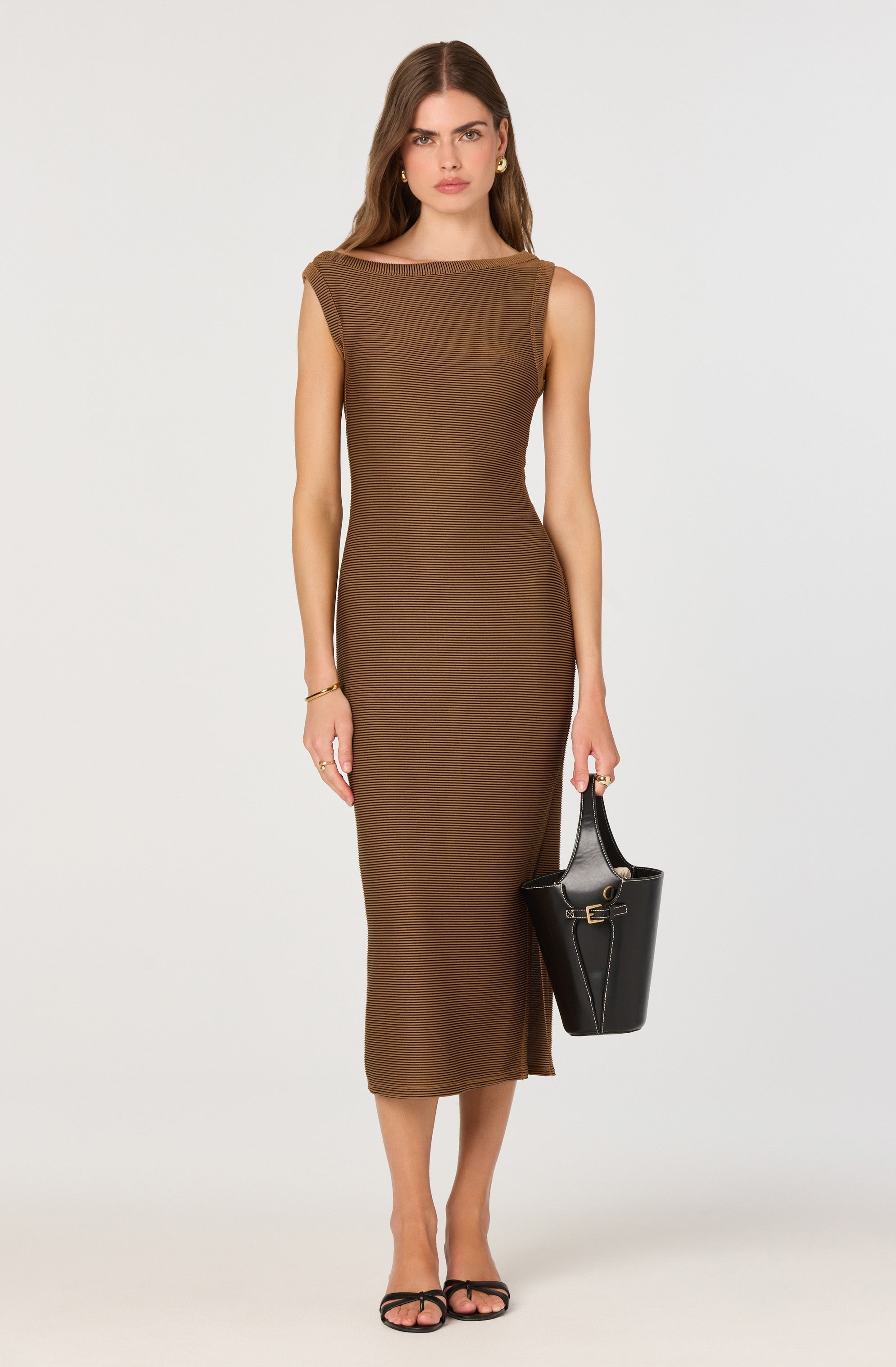 A woman is wearing a sleeveless, fitted, midi-length brown dress with a subtle ribbed texture and a high, rounded neckline. She is holding a black handbag with a gold buckle and is wearing black strappy heels.