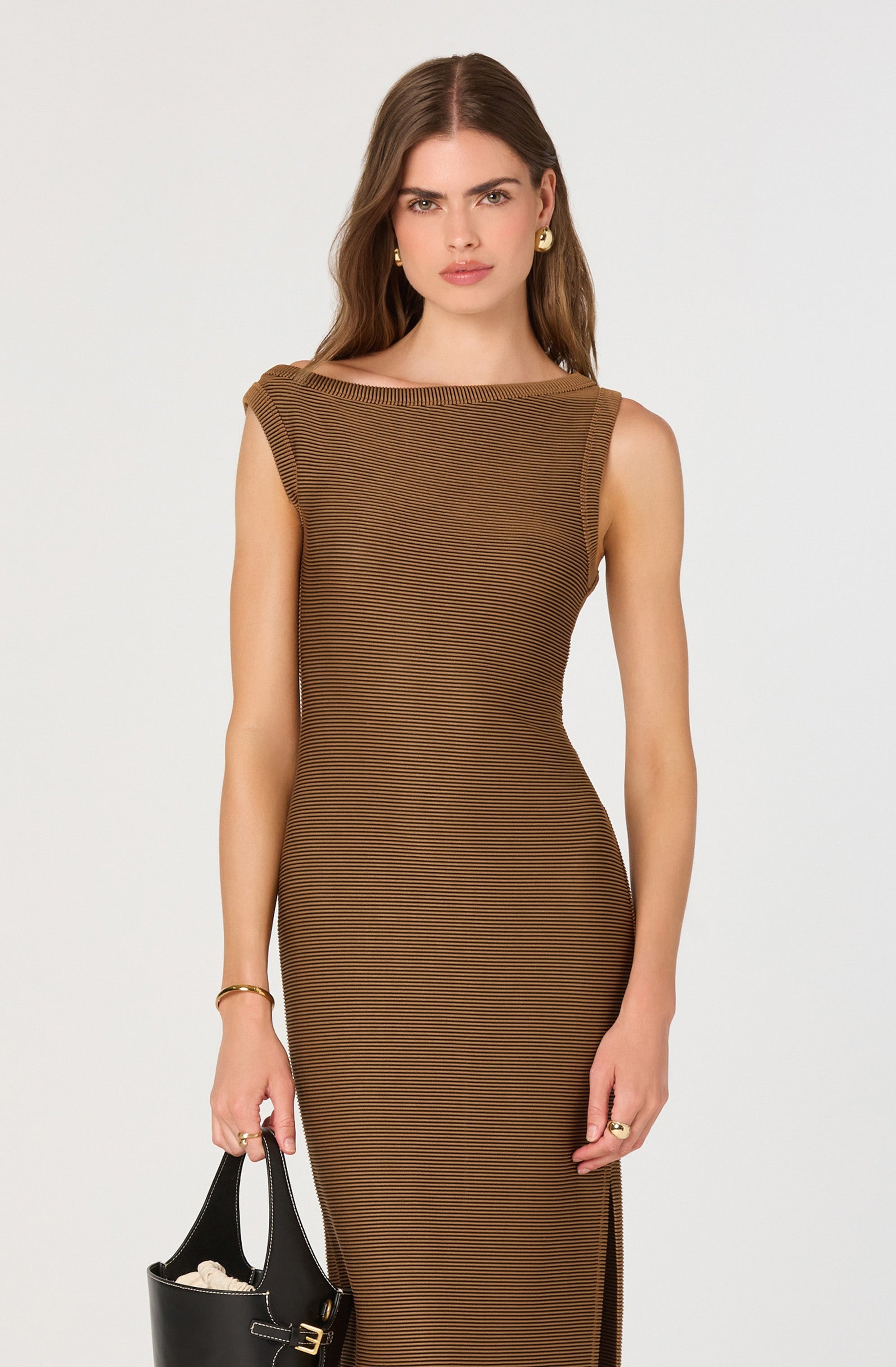A woman with long, wavy brown hair is wearing a fitted sleeveless dress with horizontal black and brown stripes. The dress has a wide, rounded neckline and a small slit on the side. She is accessorized with gold hoop earrings, a gold bracelet, and a ring on her right hand. She is holding a black handbag with a curved top edge and a small gold logo on the front.