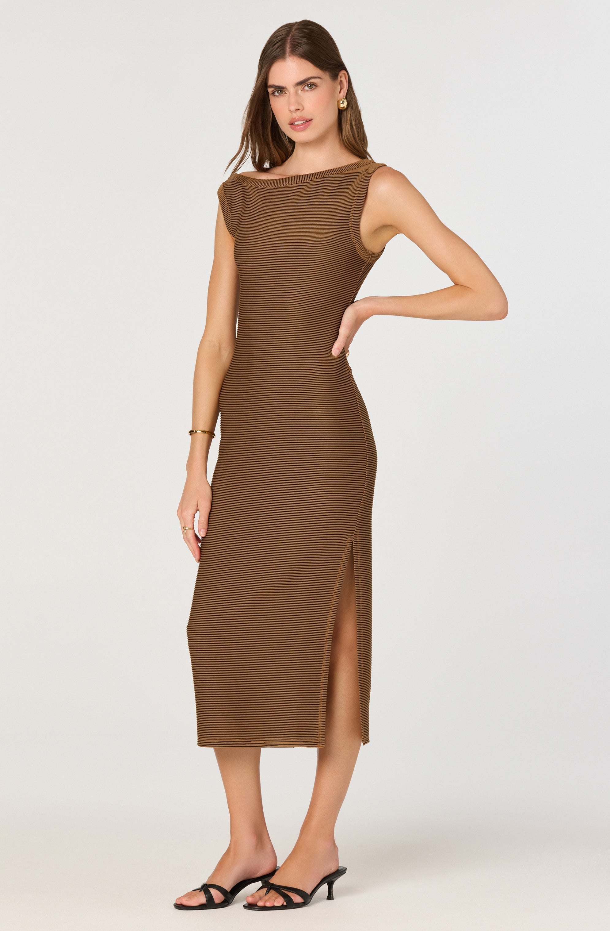 A woman is wearing a sleeveless, ankle-length dress in a brown, ribbed-knit fabric. The dress features a high, rounded neckline, and a side slit that reaches just below the knee. The fabric has a subtle horizontal stripe pattern. She is accessorized with gold earrings, a gold bracelet, and black strappy high-heeled sandals.