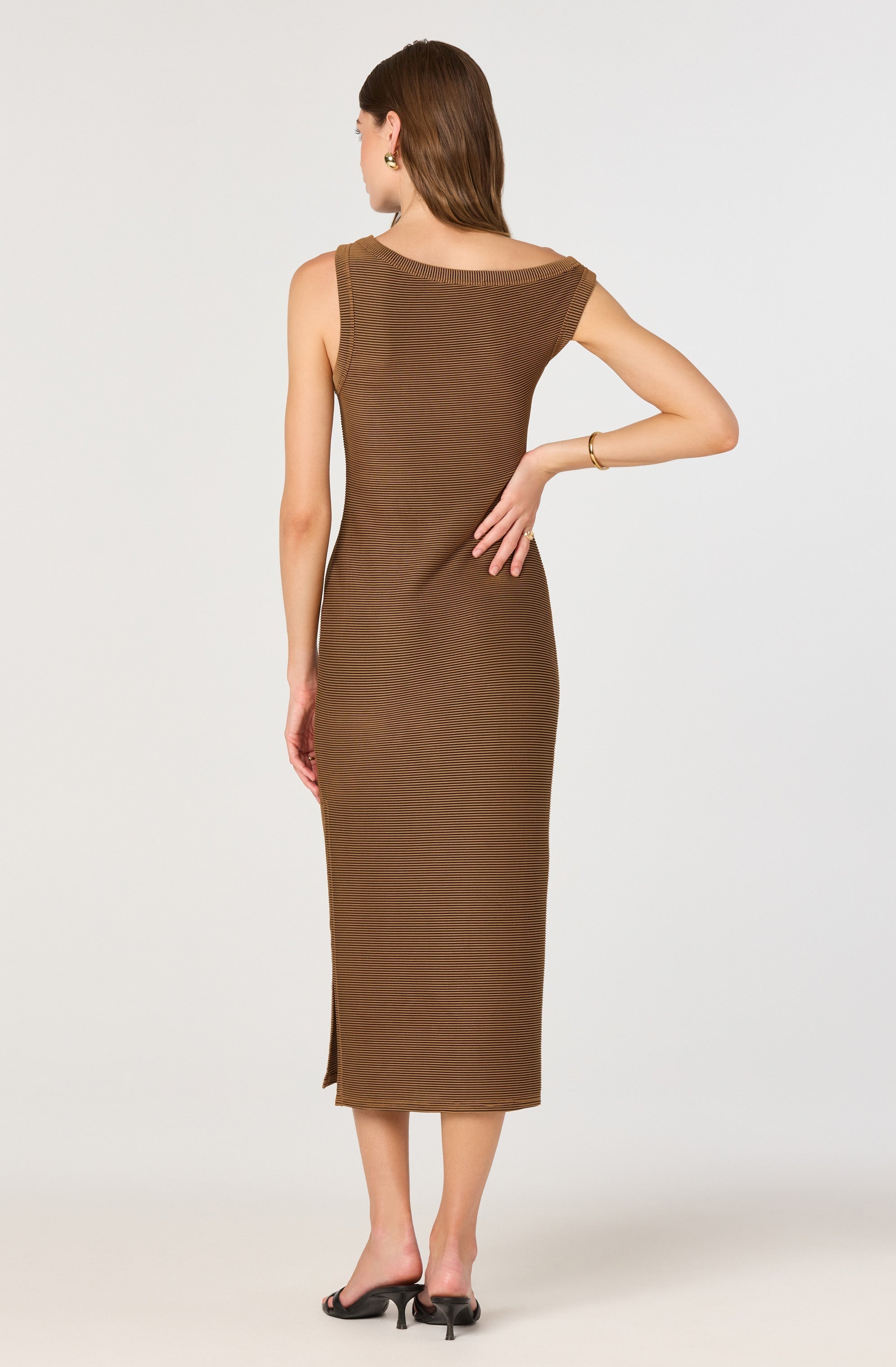 A woman is wearing a sleeveless, ankle-length brown dress with horizontal ribbed texture. The dress features a boat neckline and a side slit at the hem. She is accessorized with gold earrings and a gold bracelet on her right wrist, and is wearing black heeled sandals.