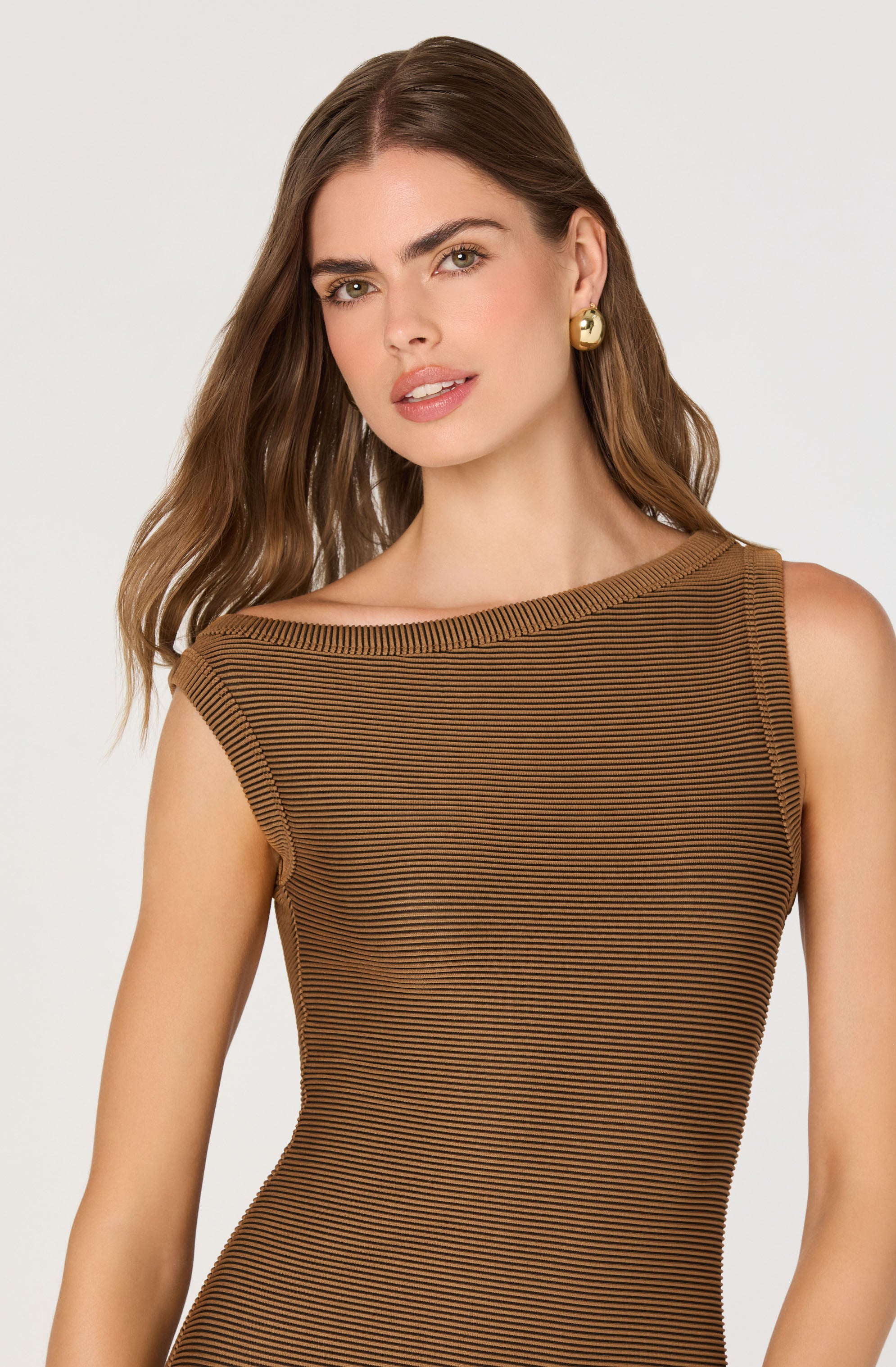 A woman is wearing a sleeveless, fitted dress with horizontal brown and black thin stripes. The dress features an asymmetrical neckline with a wide, ribbed collar detail. She has gold earrings and her hair is styled in loose waves.