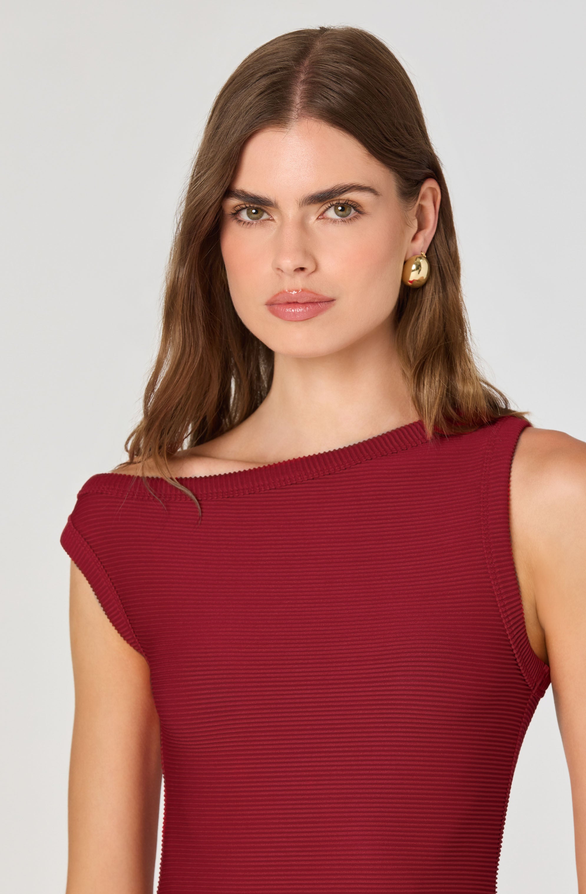 A woman is wearing a sleeveless, form-fitting dress in a rich burgundy color. The dress has a subtle, fine horizontal ribbed texture. She is accessorized with large, gold, round earrings.
